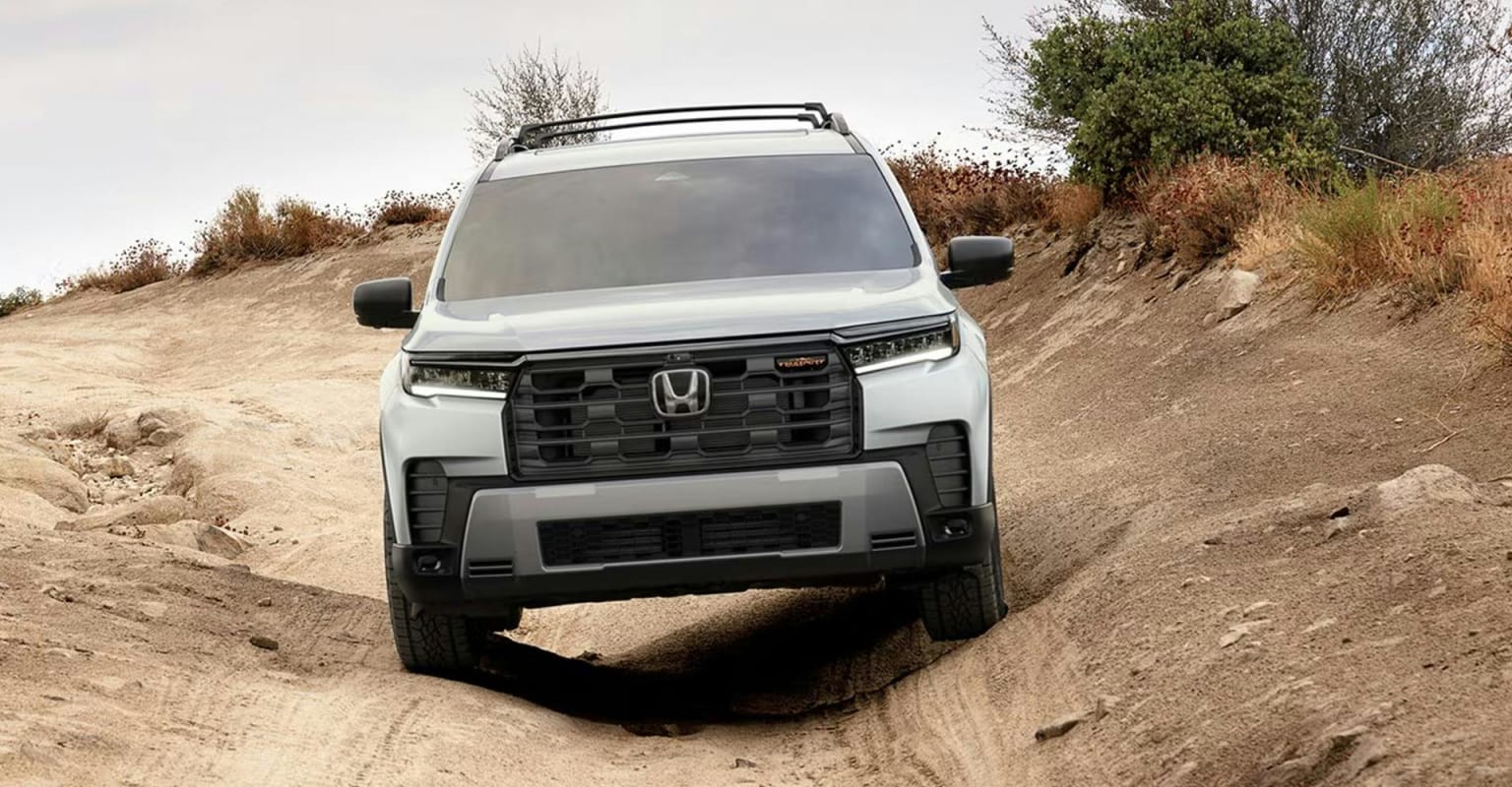A white SUV navigates a rocky, dirt path with sparse vegetation in the background.