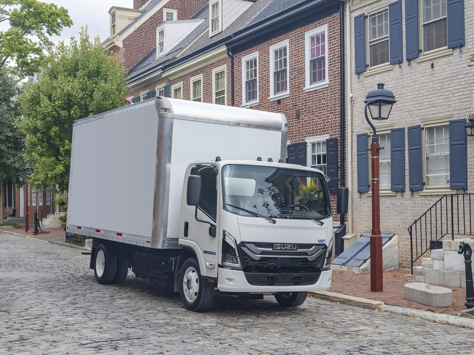 A white box truck parked on a cobblestone street in front of historic brick buildings with blue shutters.