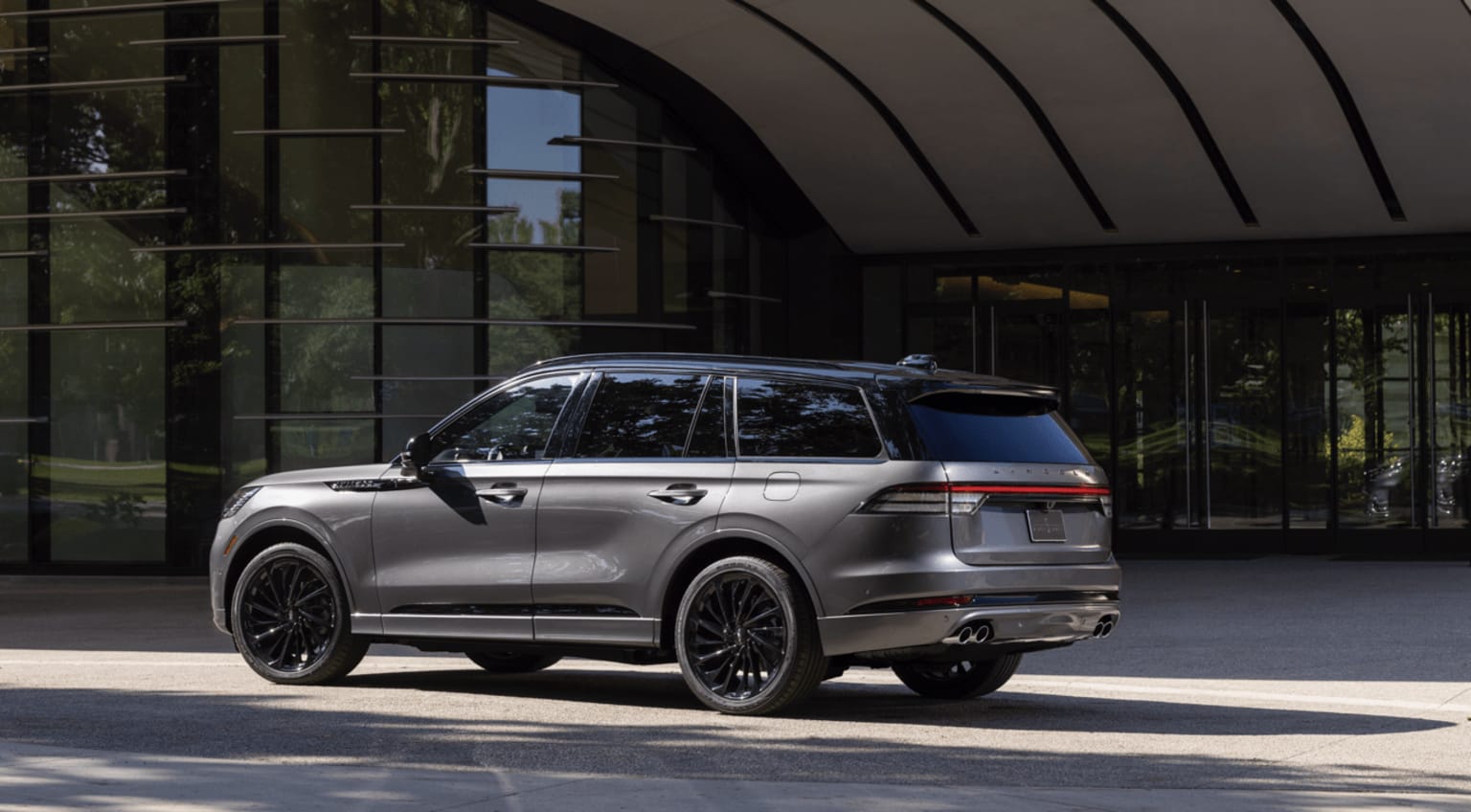A sleek, silver SUV parked in front of a modern, glass-enclosed building, with bold design and dark accents creating a striking contrast against the lush, green surroundings.