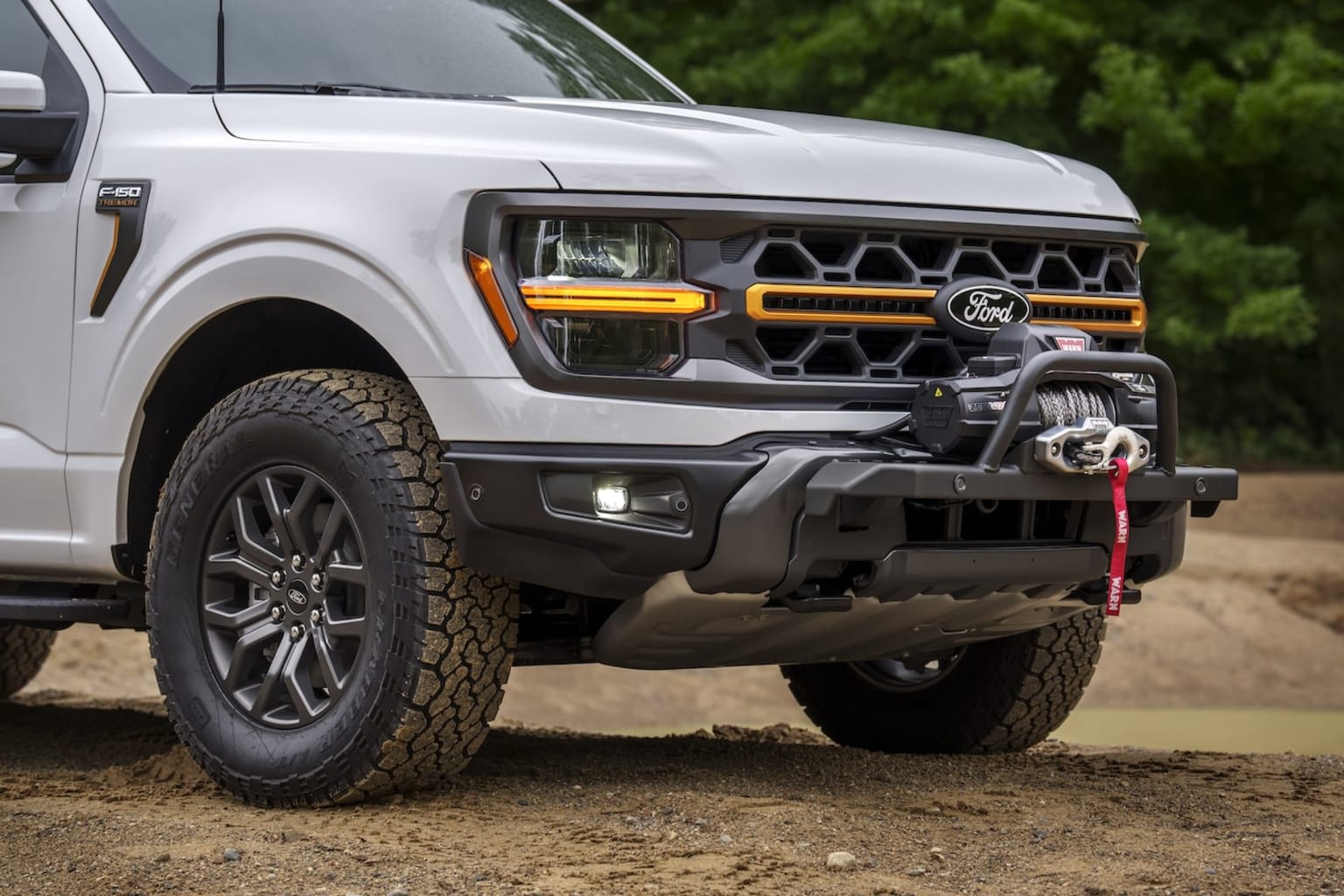 An off-road-ready pickup truck with large tires and a bold, aggressive front grille stands on a dirt surface surrounded by lush greenery.