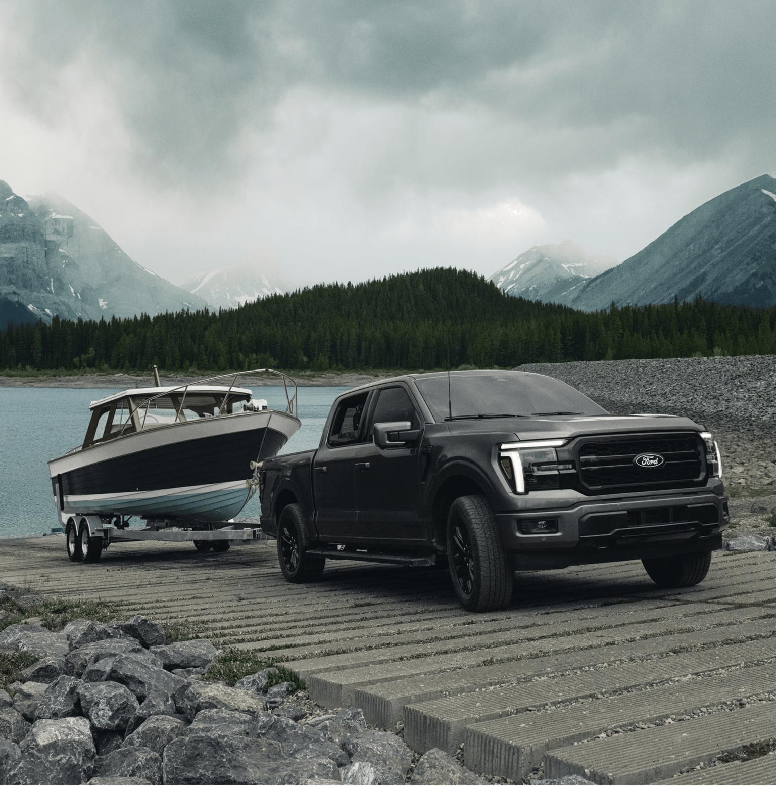 A black pickup truck towing a boat on a trailer sits on a concrete ramp with a lake, evergreen forests, and misty mountains in the background.