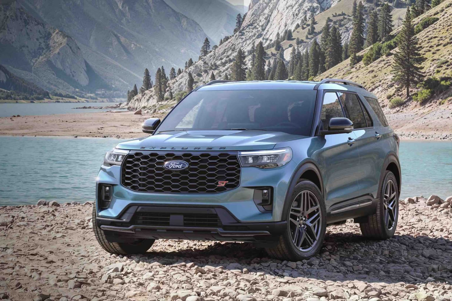A teal SUV sits on a rocky shore with a calm lake and mountains in the background.
