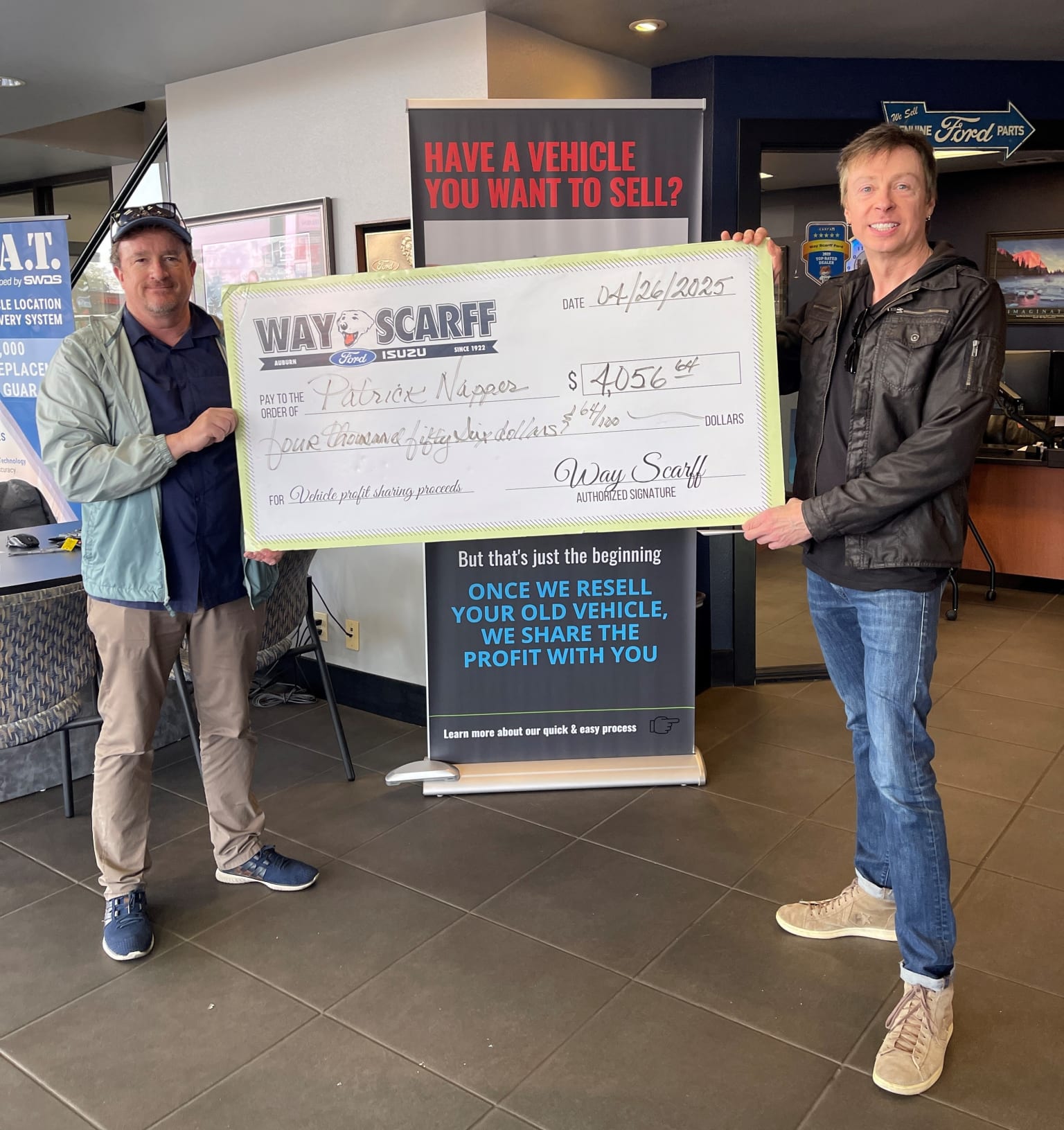 Two men holding a large check in front of a car dealership, with a sign advertising the sale of vehicles.