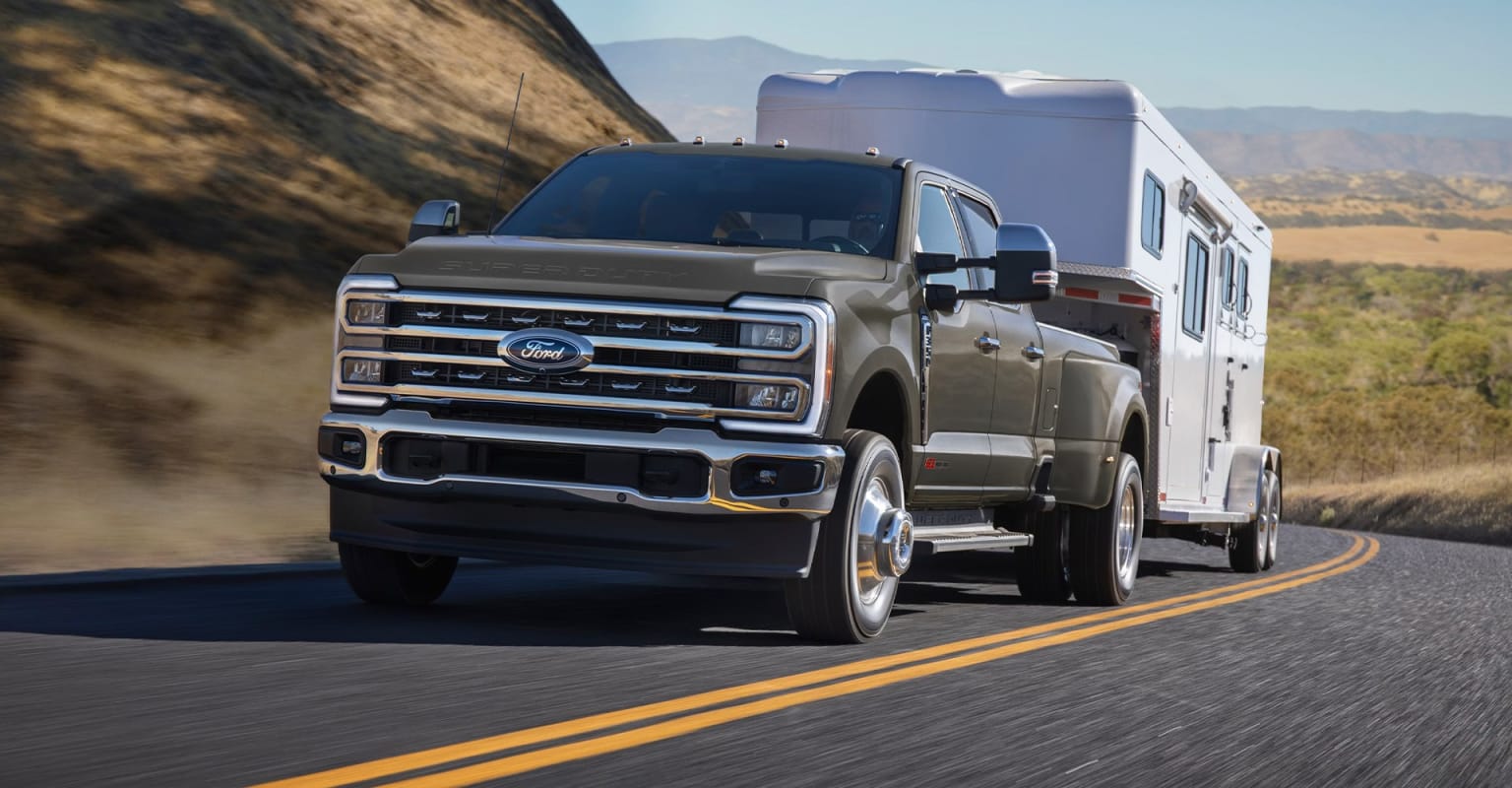 A large, rugged pickup truck towing a recreational vehicle on a winding road surrounded by mountains and a clear sky.