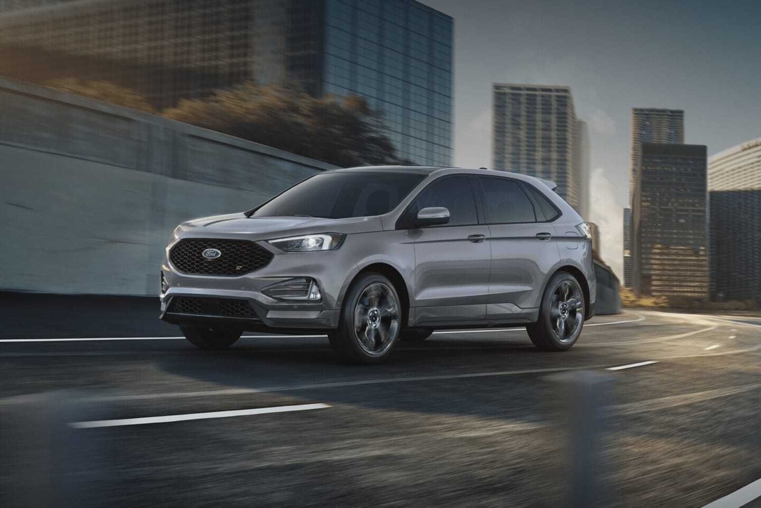 A sleek, silver SUV driving along a city street with glass skyscrapers in the background.