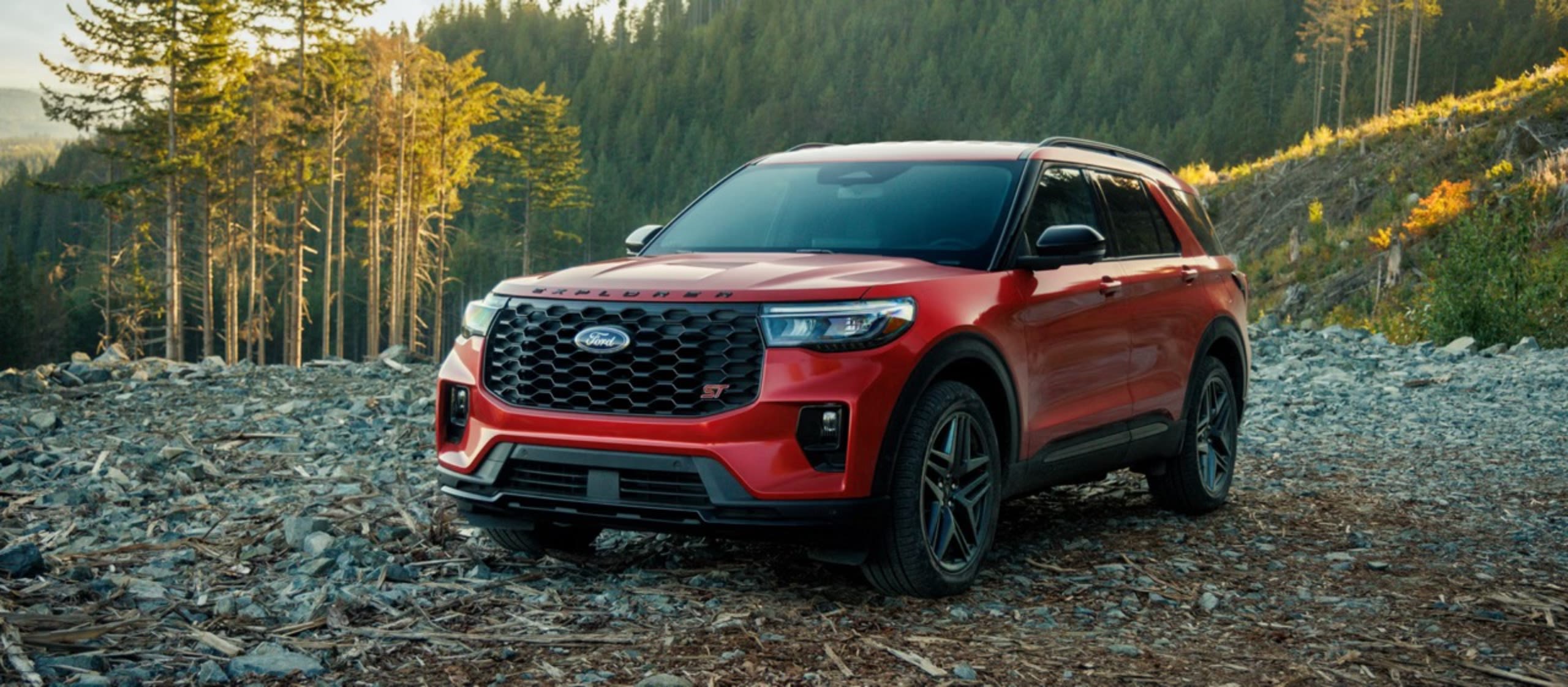 A red SUV sits on rocky terrain with a dense evergreen forest in the background.