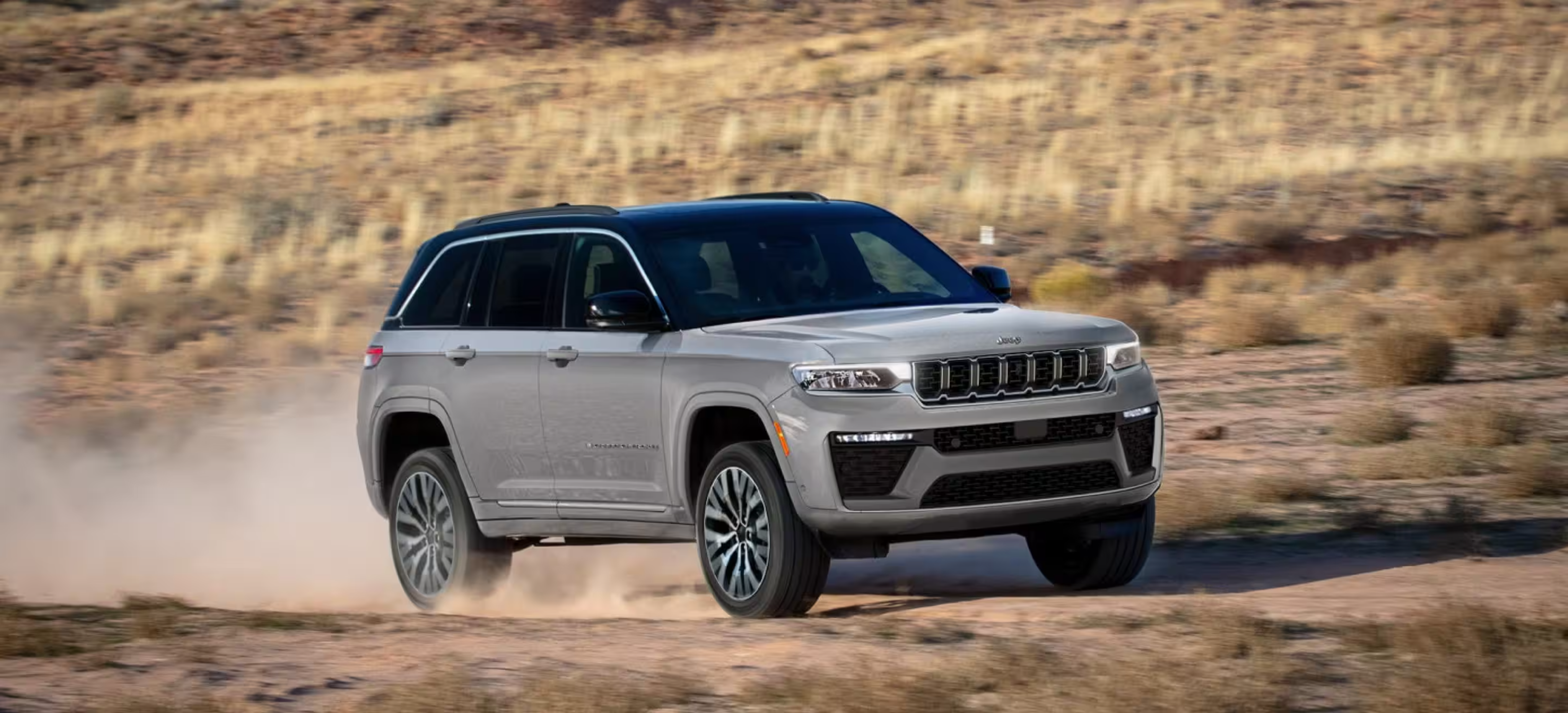 A silver SUV drives through a dusty desert landscape with dry brush and rolling hills in the background.