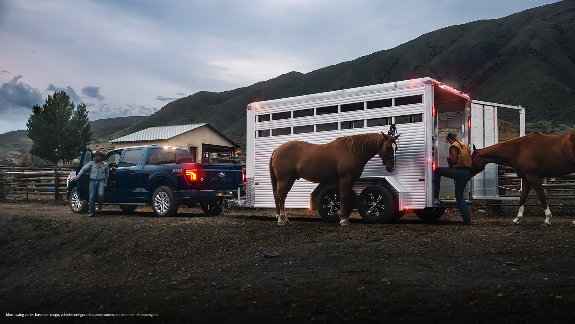 A dark blue pickup truck is hitched to a horse trailer, with two horses being loaded into it, set against a backdrop of rolling hills and a cloudy sky.