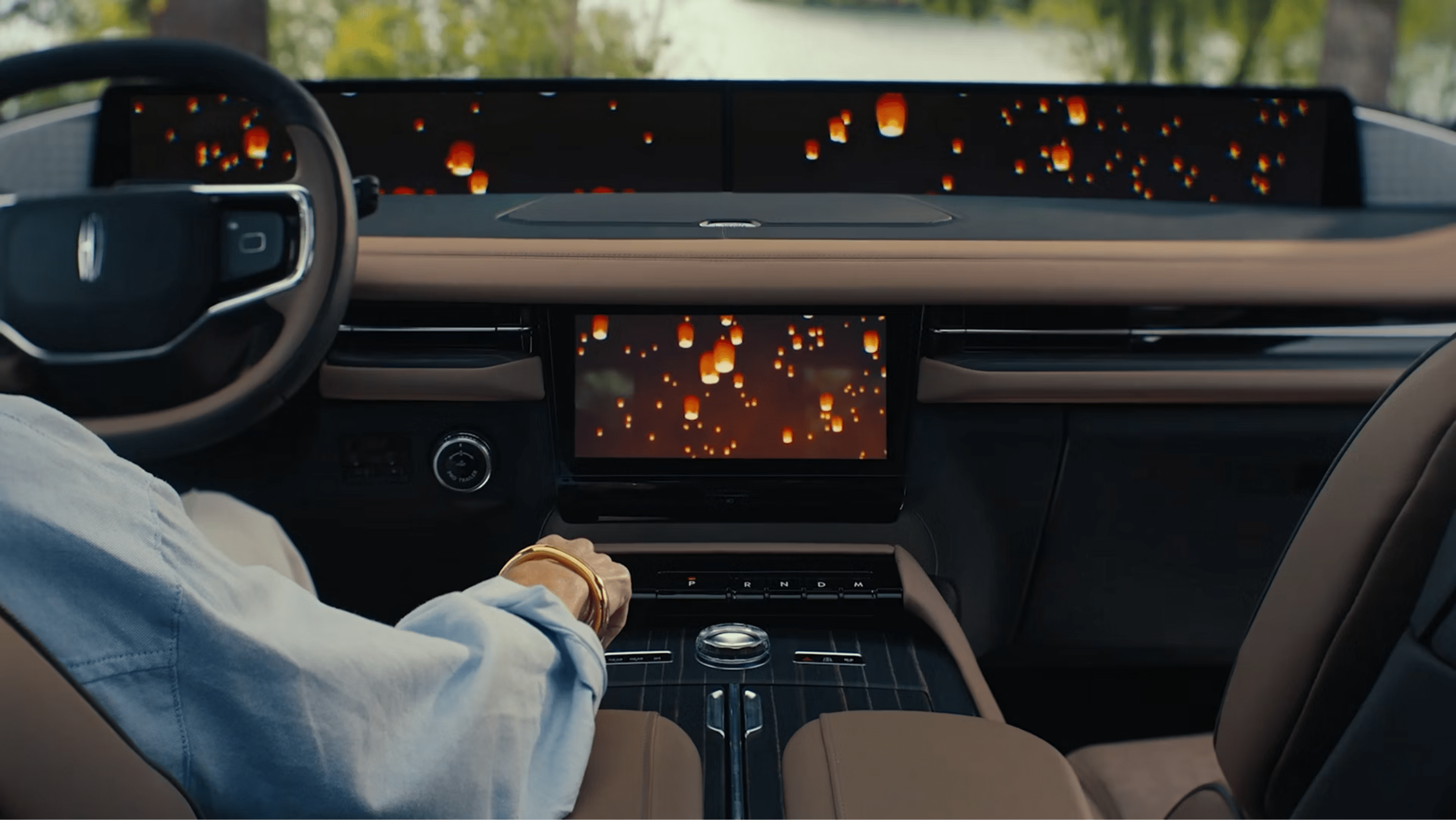 A person's arm rests on the center console of a car interior with multiple digital screens showing floating lanterns, and a blurred natural landscape visible through the windshield.