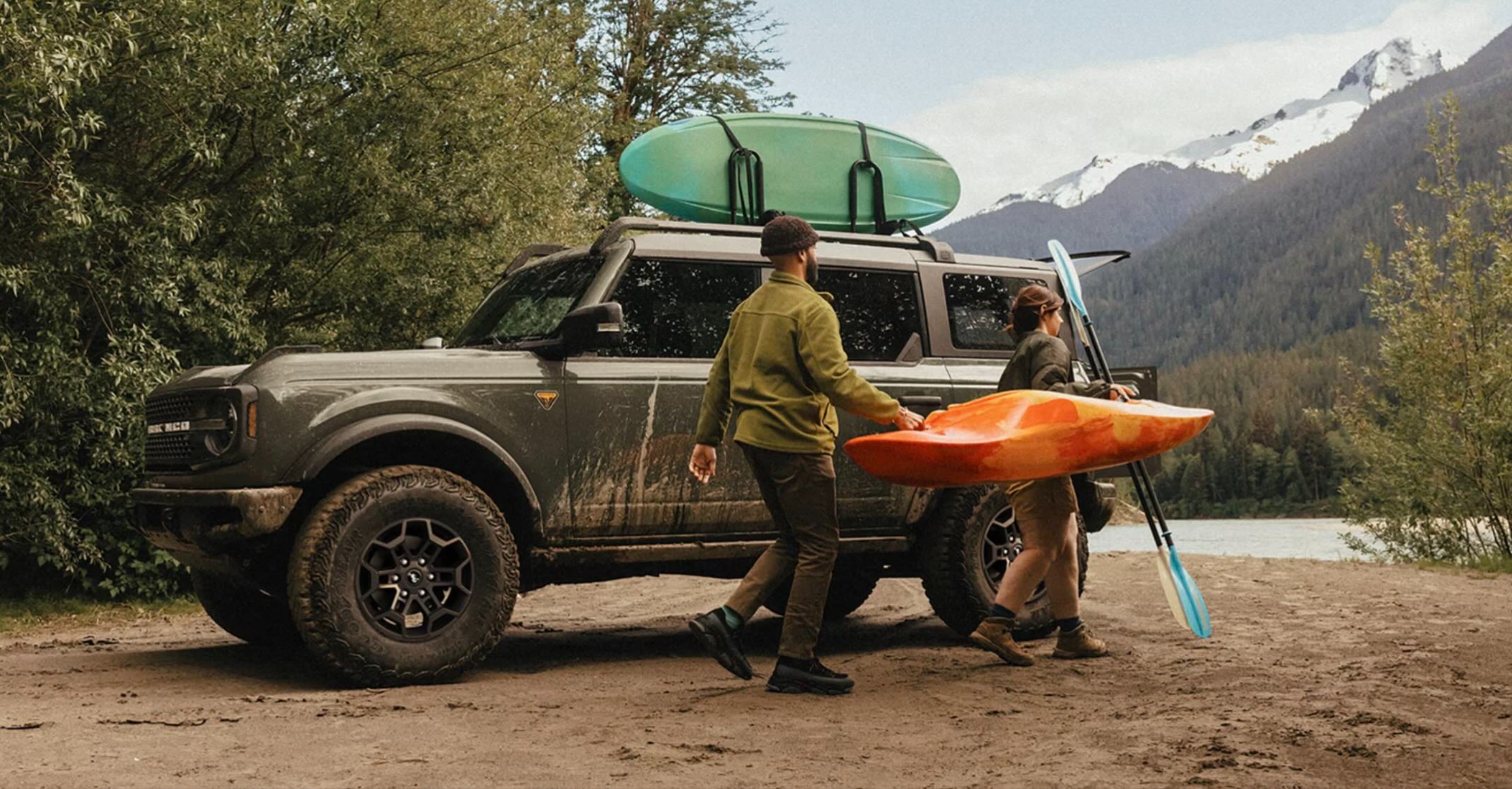 Two people carry a kayak and paddles past a rugged SUV loaded with outdoor gear, with a serene lake and snow-capped mountains in the background.