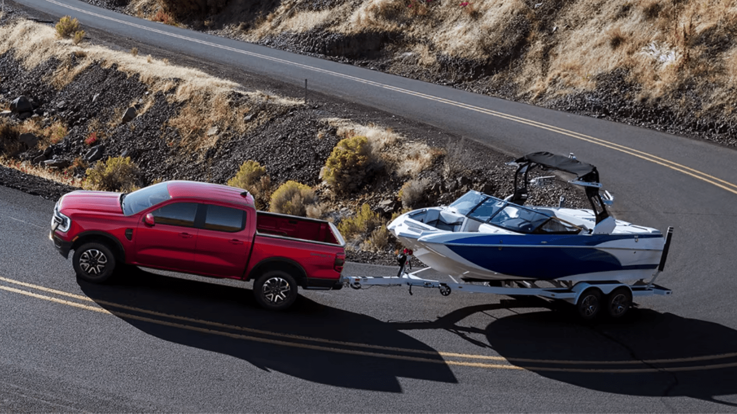 A red pickup truck towing a blue and white boat on a trailer along a winding road with a rocky, arid hillside in the background.
