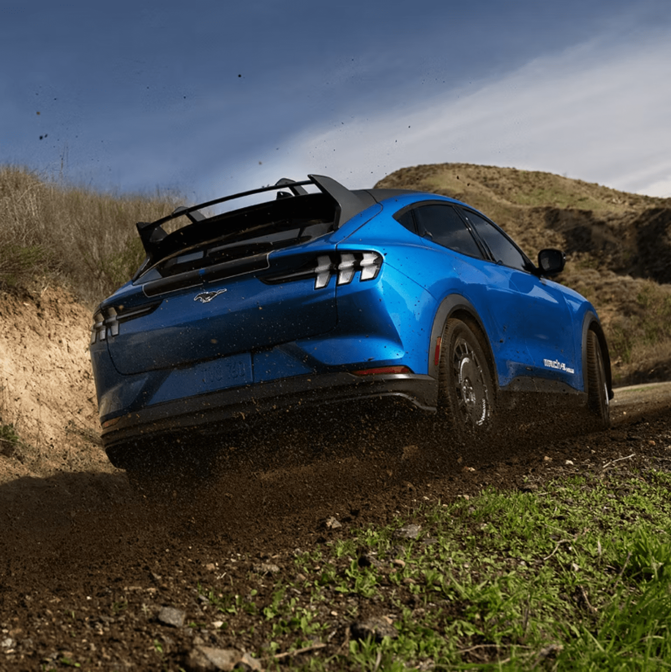 A blue electric SUV kicks up dirt as it drives on a gravel road with rolling hills and a cloudy sky in the background.