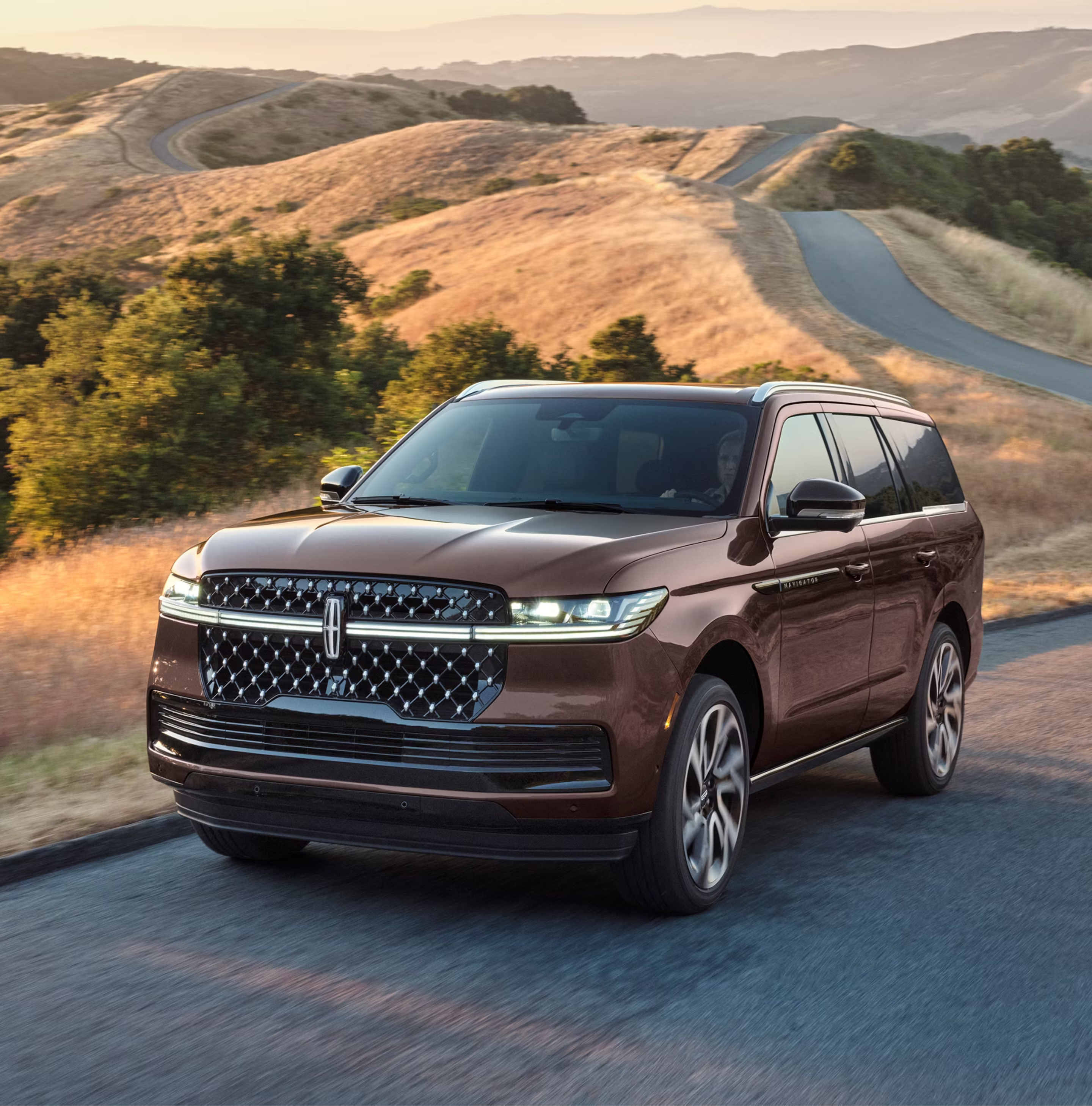 A brown SUV drives along a winding road through rolling golden hills under a soft sunset sky.