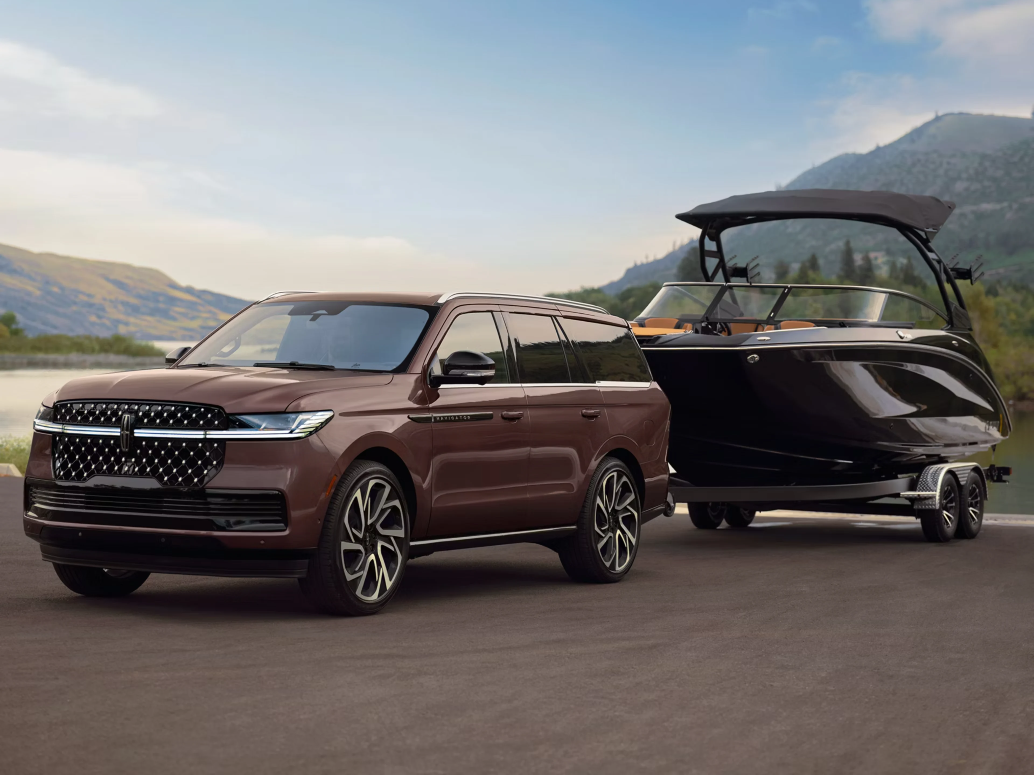 A brown luxury SUV towing a black boat on a trailer by a serene lake with mountains in the background.