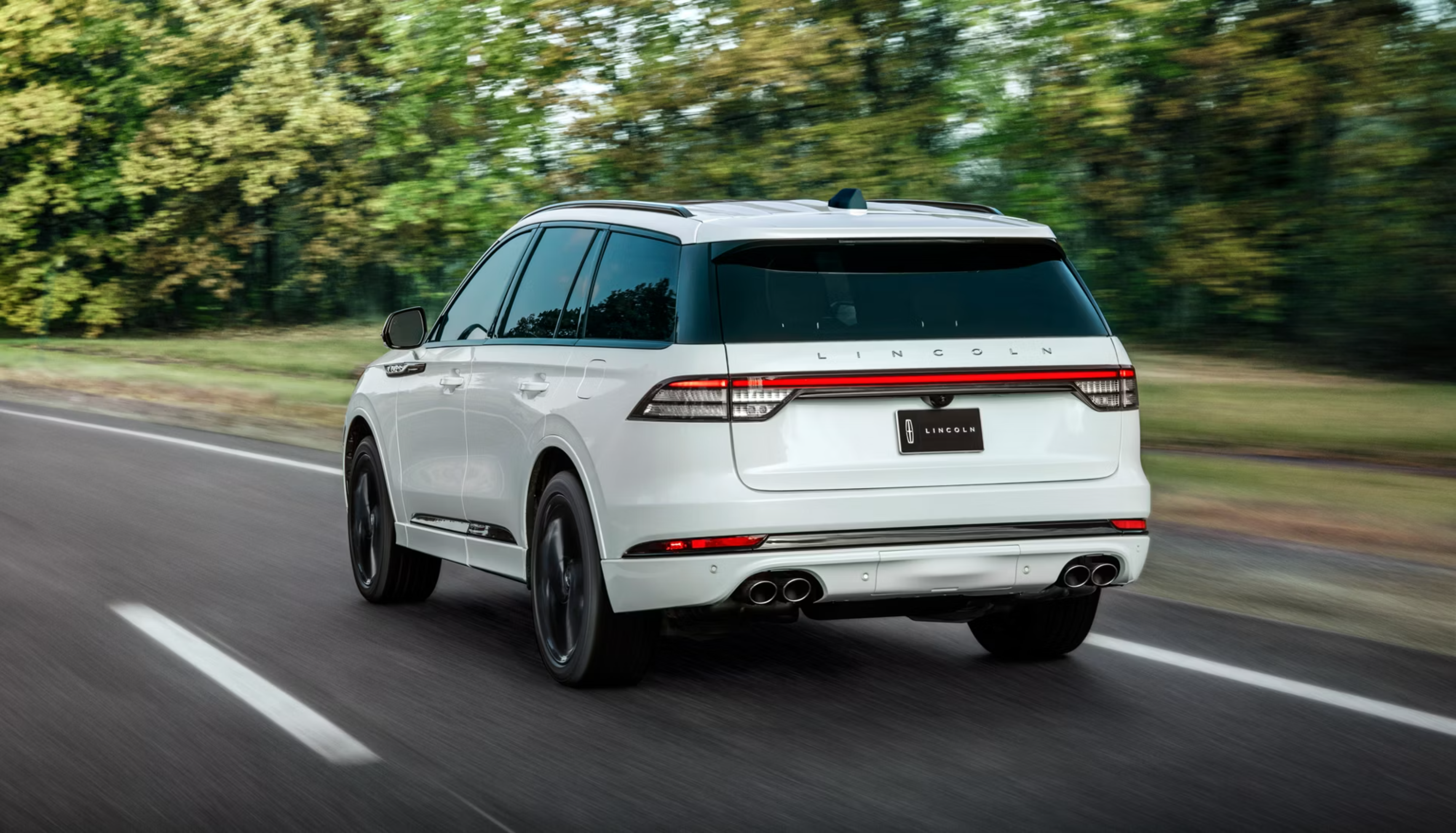 A white SUV drives down a road with trees and foliage in the background.