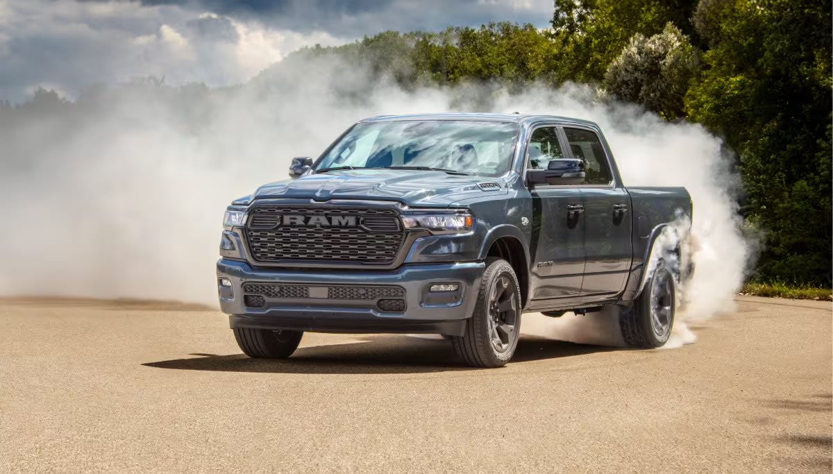 A grey pickup truck performs a burnout on a paved road with white smoke, trees, and cloudy sky in the background.
