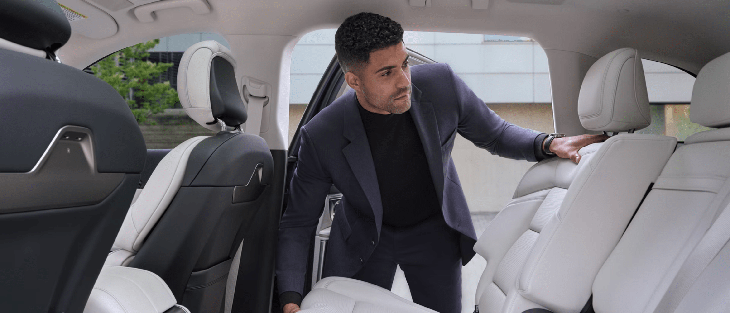 A man in a dark suit leans into the white leather interior of a car.