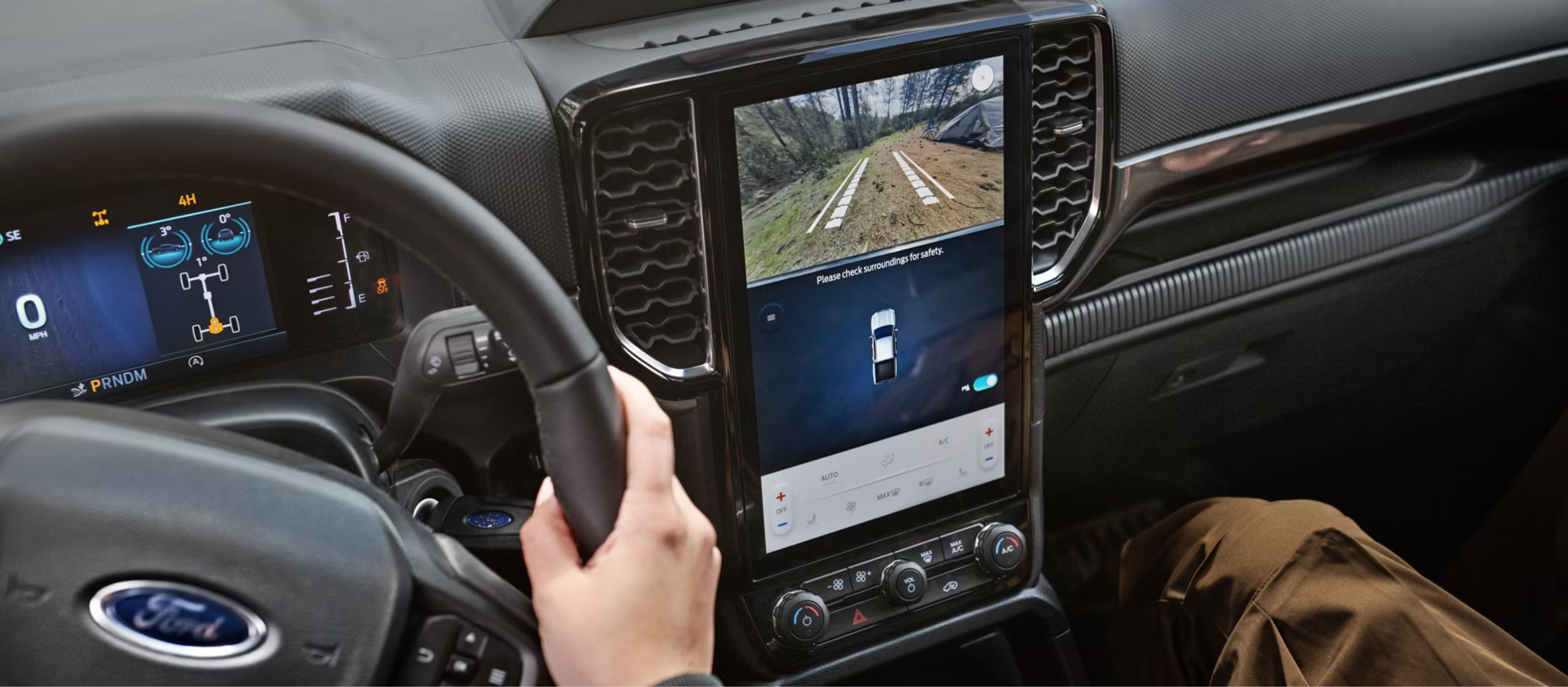A hand grips the steering wheel of a vehicle in the foreground, while a large center display shows a backup camera view against the car's interior dashboard background.