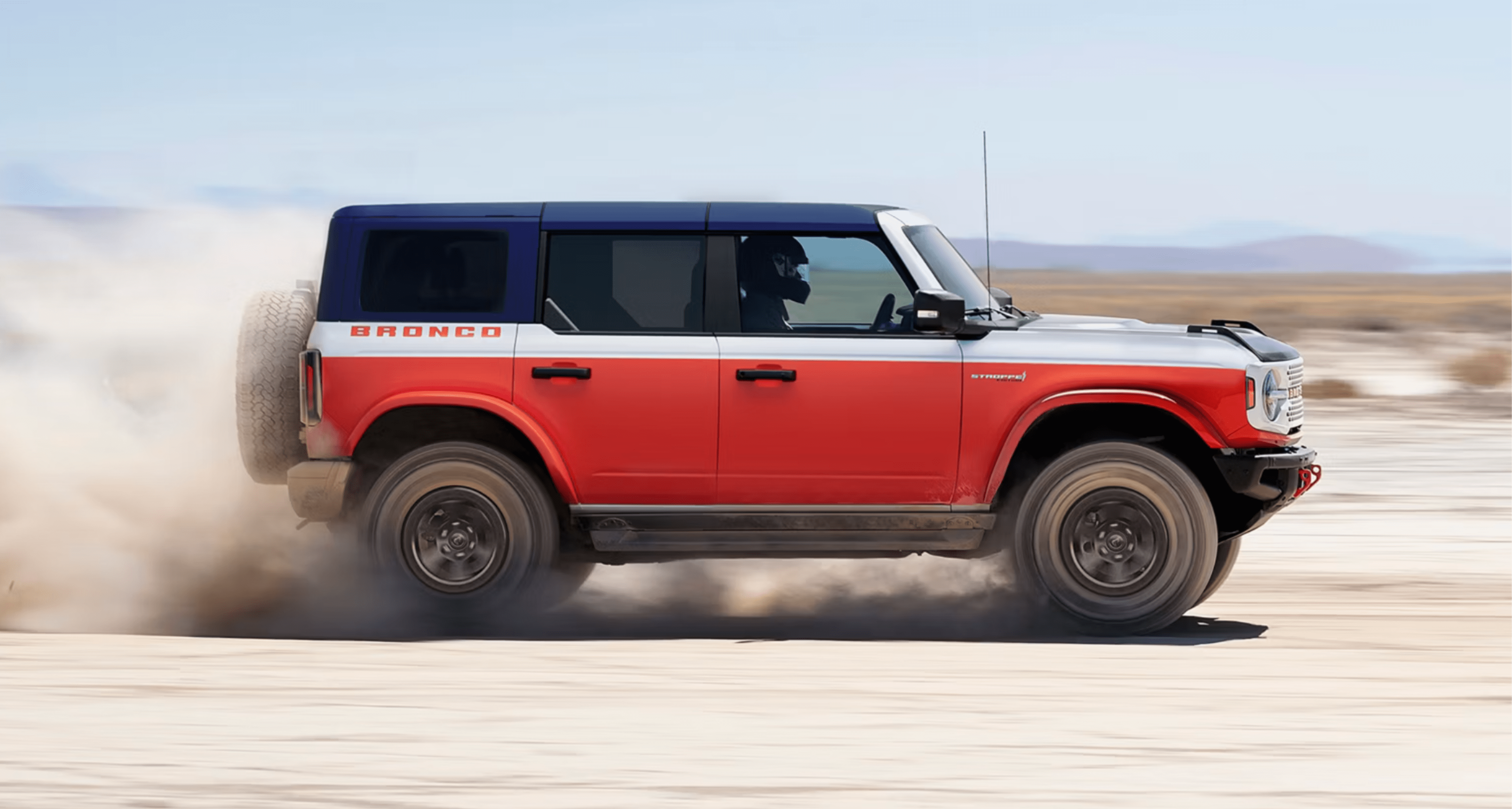 A red, white, and blue SUV kicks up dust as it drives across a dry, desert landscape under a clear sky.