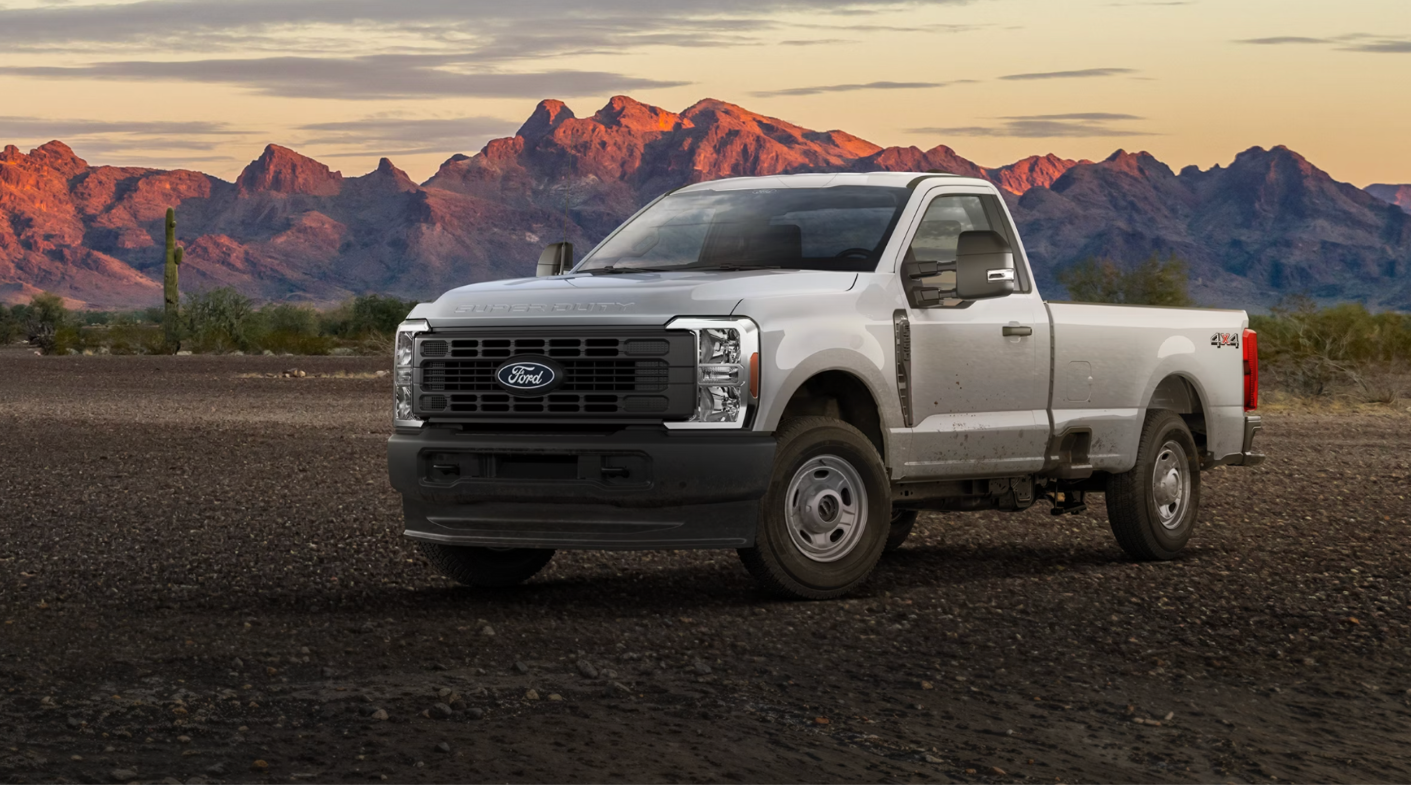 A white pickup truck sits on a gravel plain with mountains in the background.