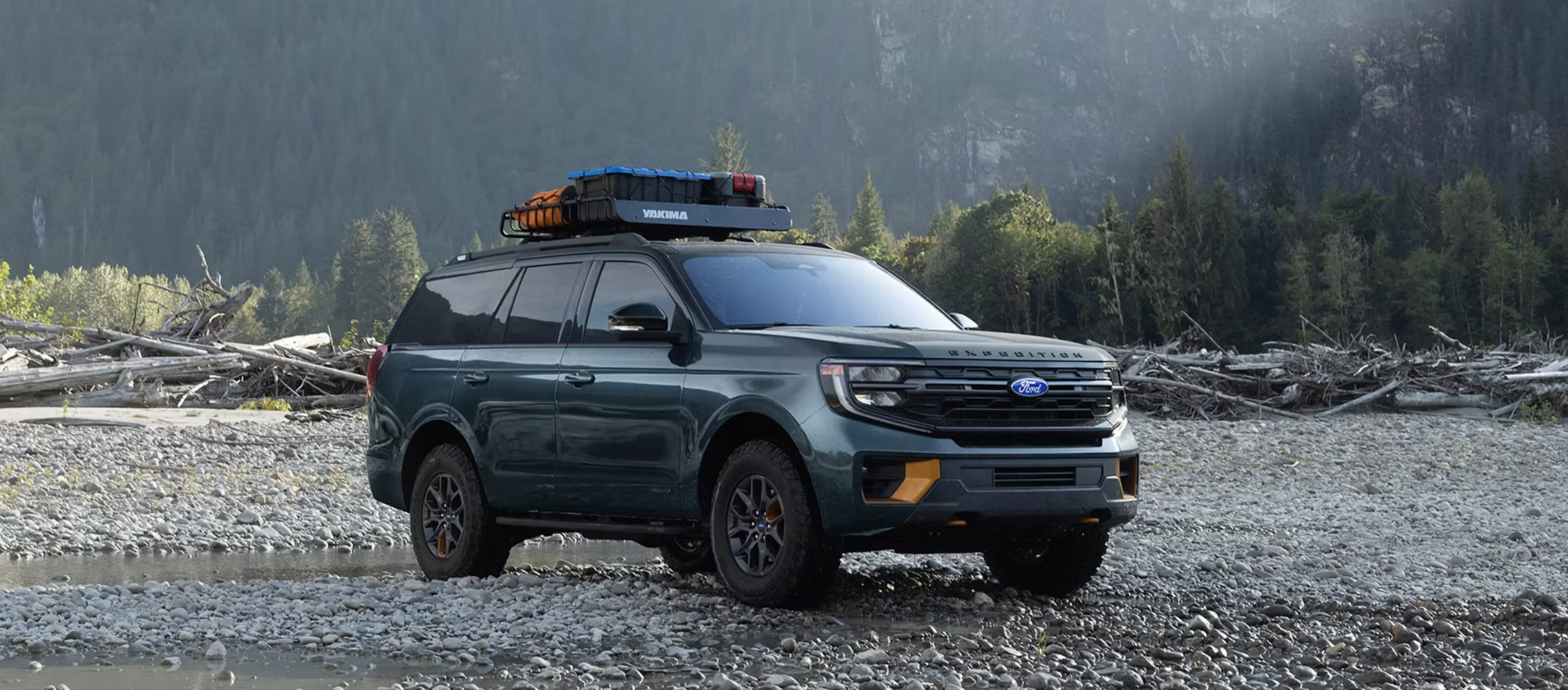 A dark green full-size SUV with a roof rack sits on a rocky riverbed against a backdrop of dense evergreen forests and misty mountains.