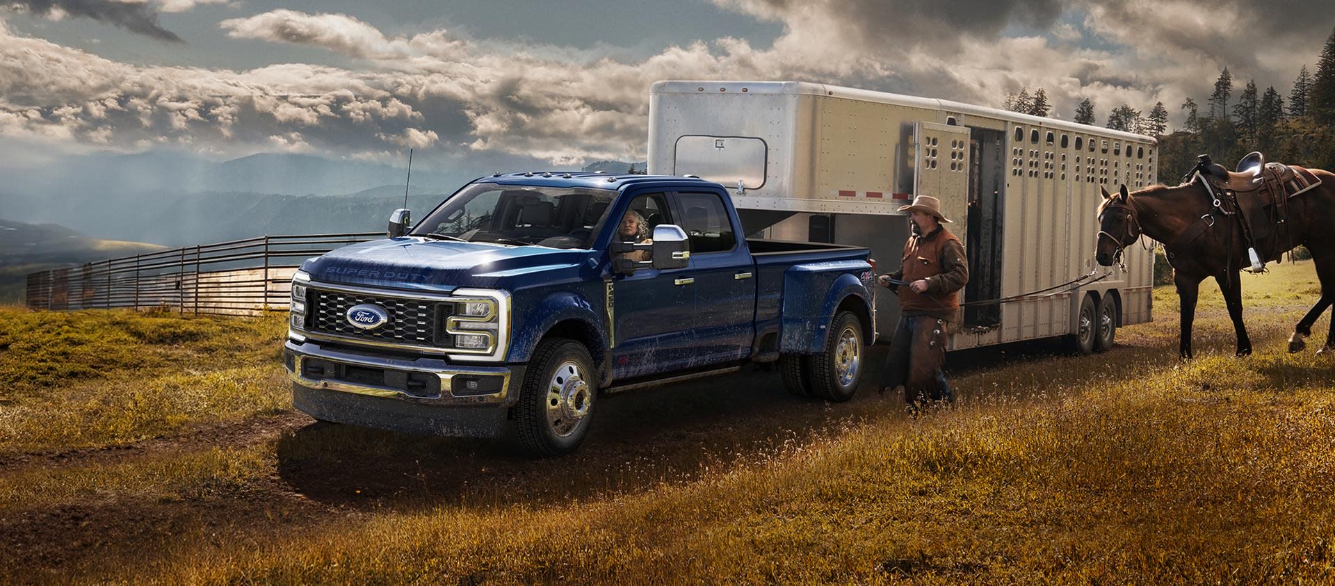 A blue pickup truck towing a large trailer driving through a grassy field with horses in the background and dramatic cloudy skies overhead.