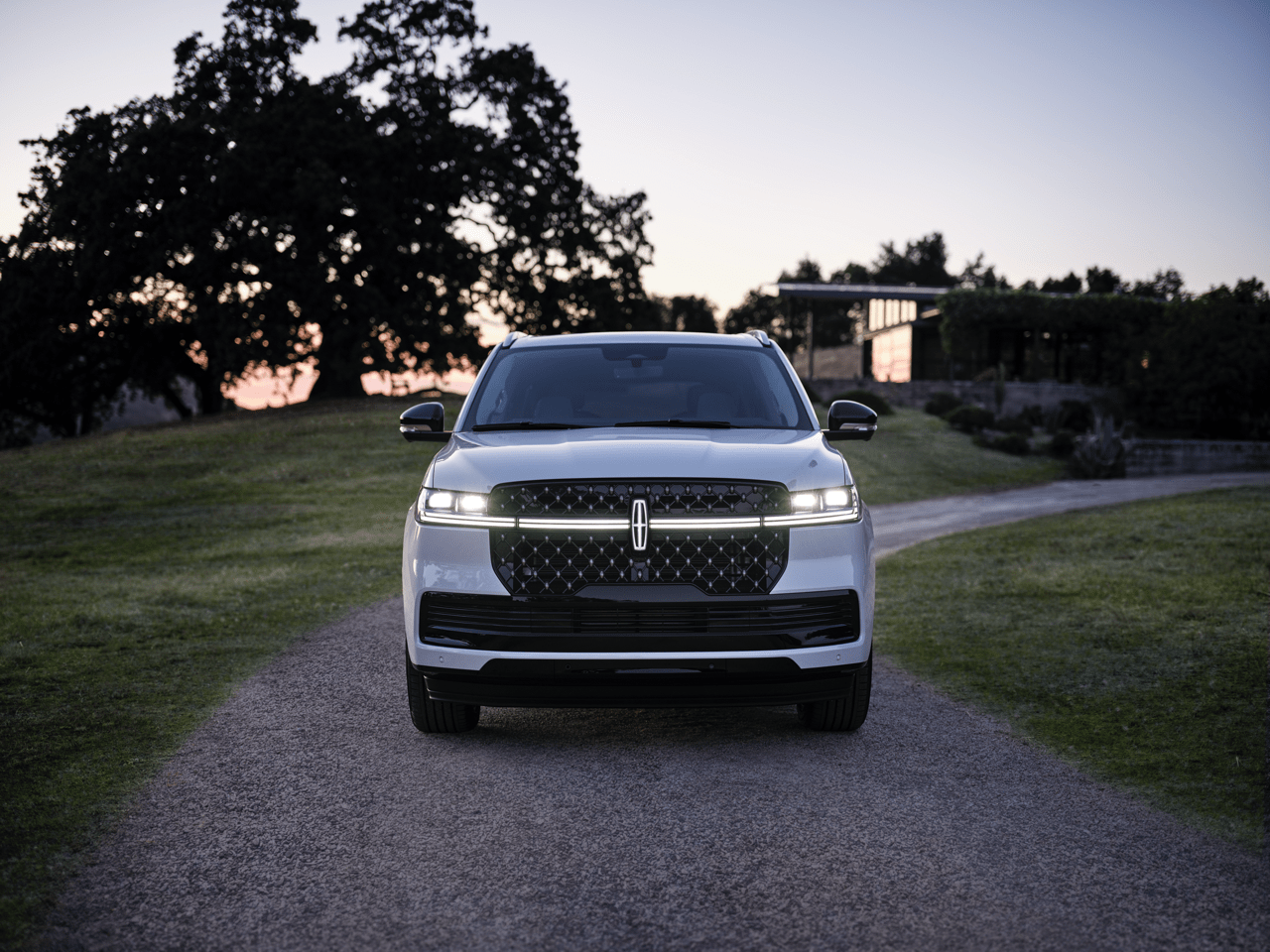 Large silver SUV in a gravel driveway with lush greenery and a sunset sky in the background