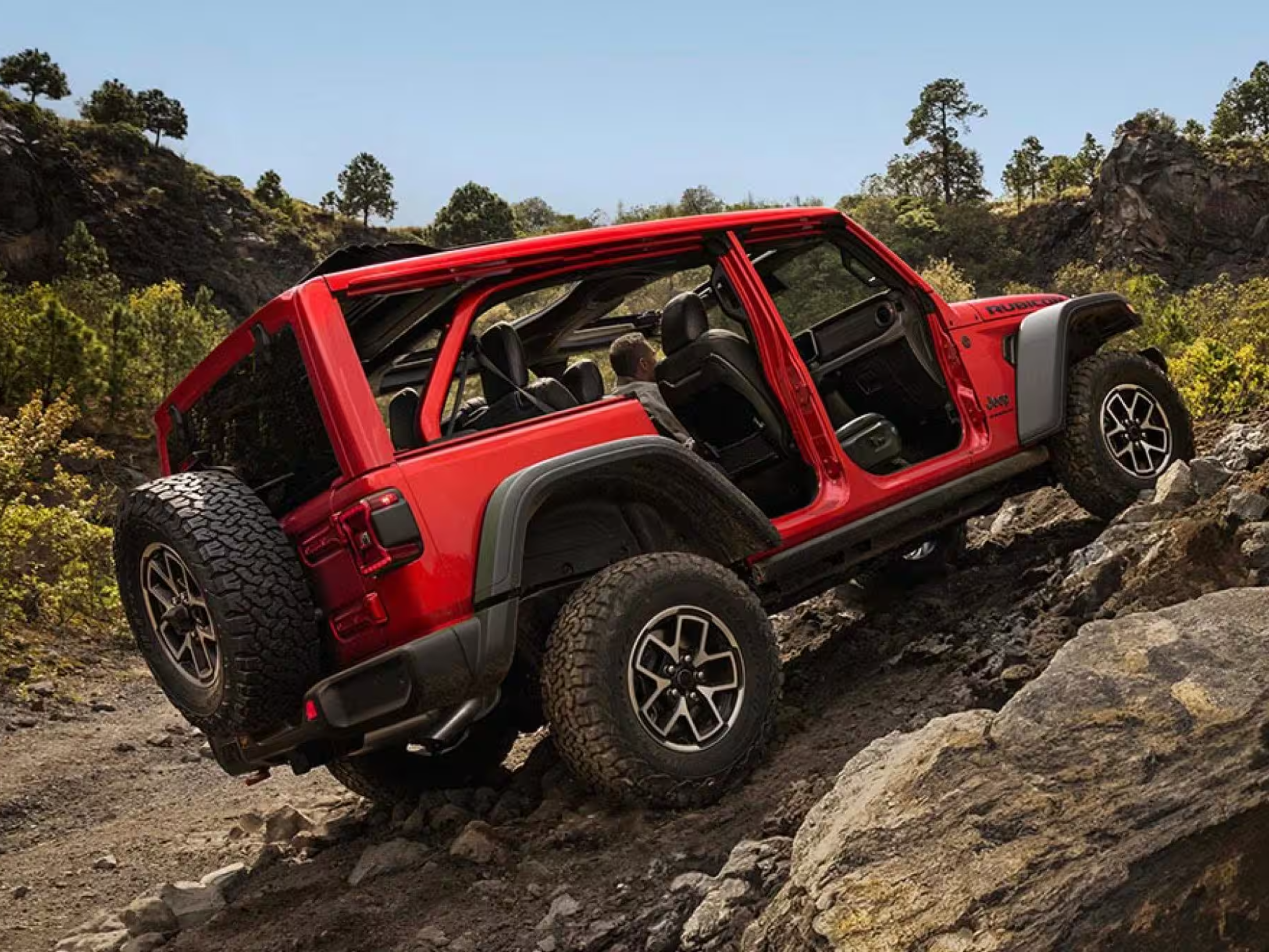 A red open-top SUV climbs a rocky incline against a backdrop of sparse trees and a clear blue sky.