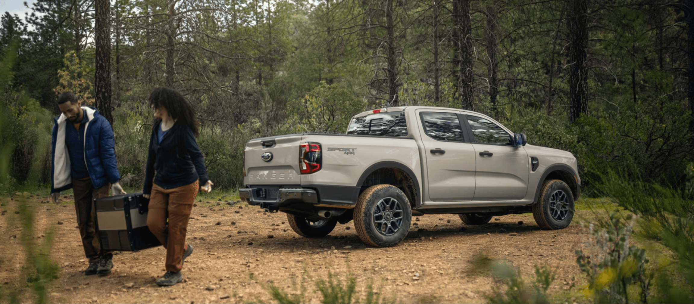 Two people carrying a large black case next to a tan pickup truck parked on a dirt path in a dense forest.