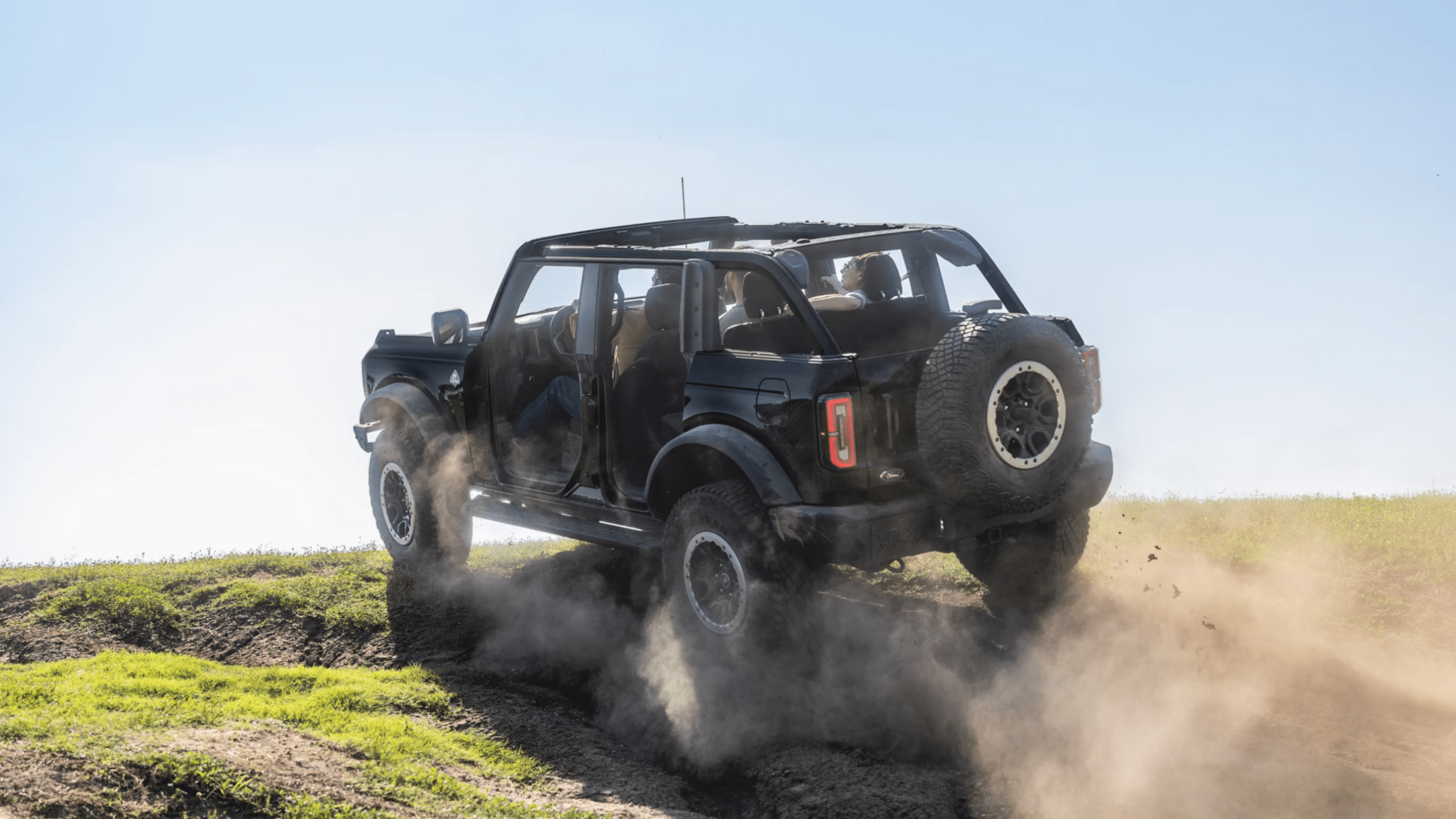A rugged off-road vehicle kicks up clouds of dust while driving across a grassy hill under a clear blue sky.