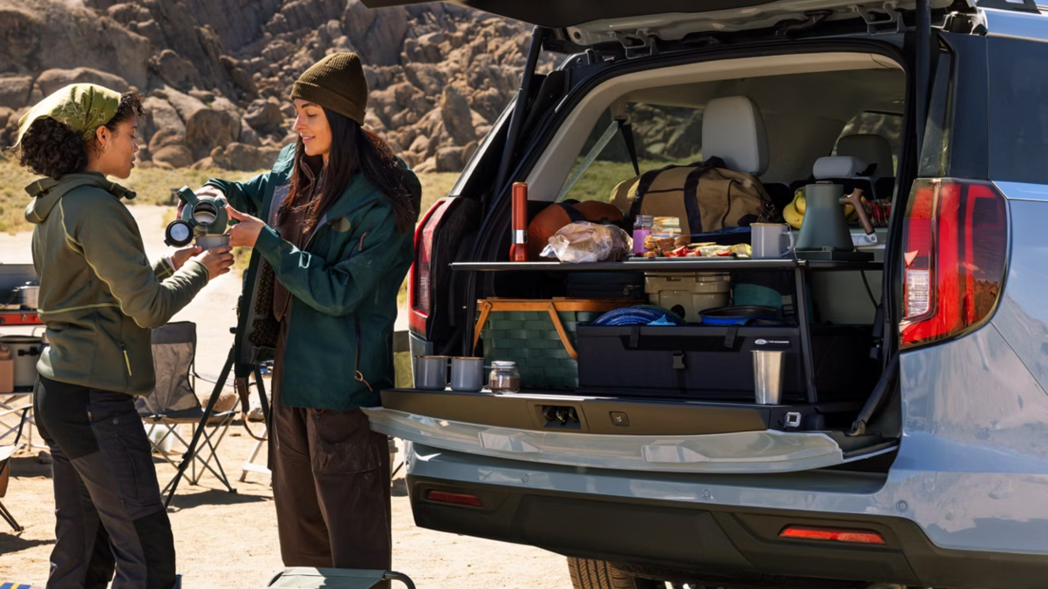 Two women stand by the open trunk of a silver SUV filled with camping gear, set against a backdrop of rugged, rocky mountains.