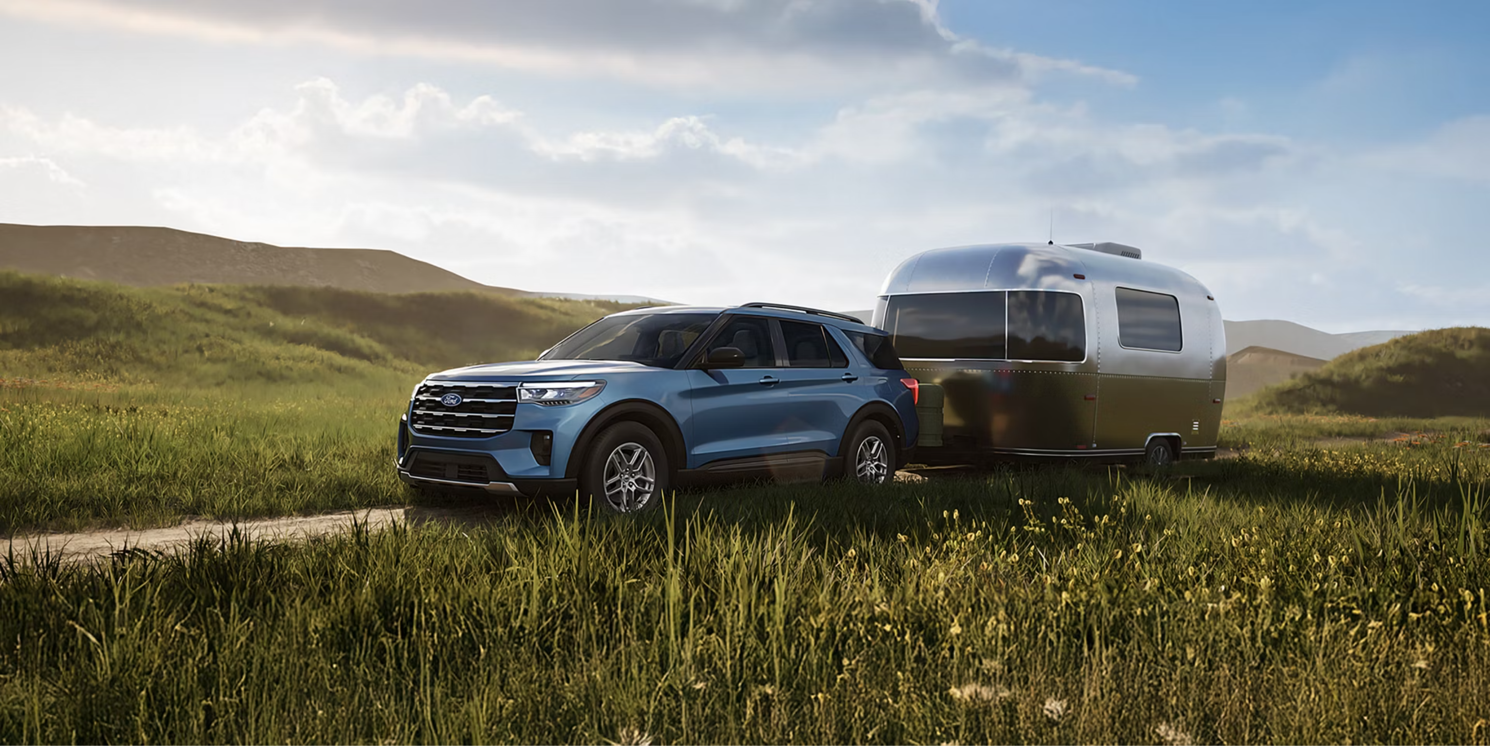 A blue SUV towing a silver travel trailer in a grassy field with rolling hills and a cloudy sky in the background.