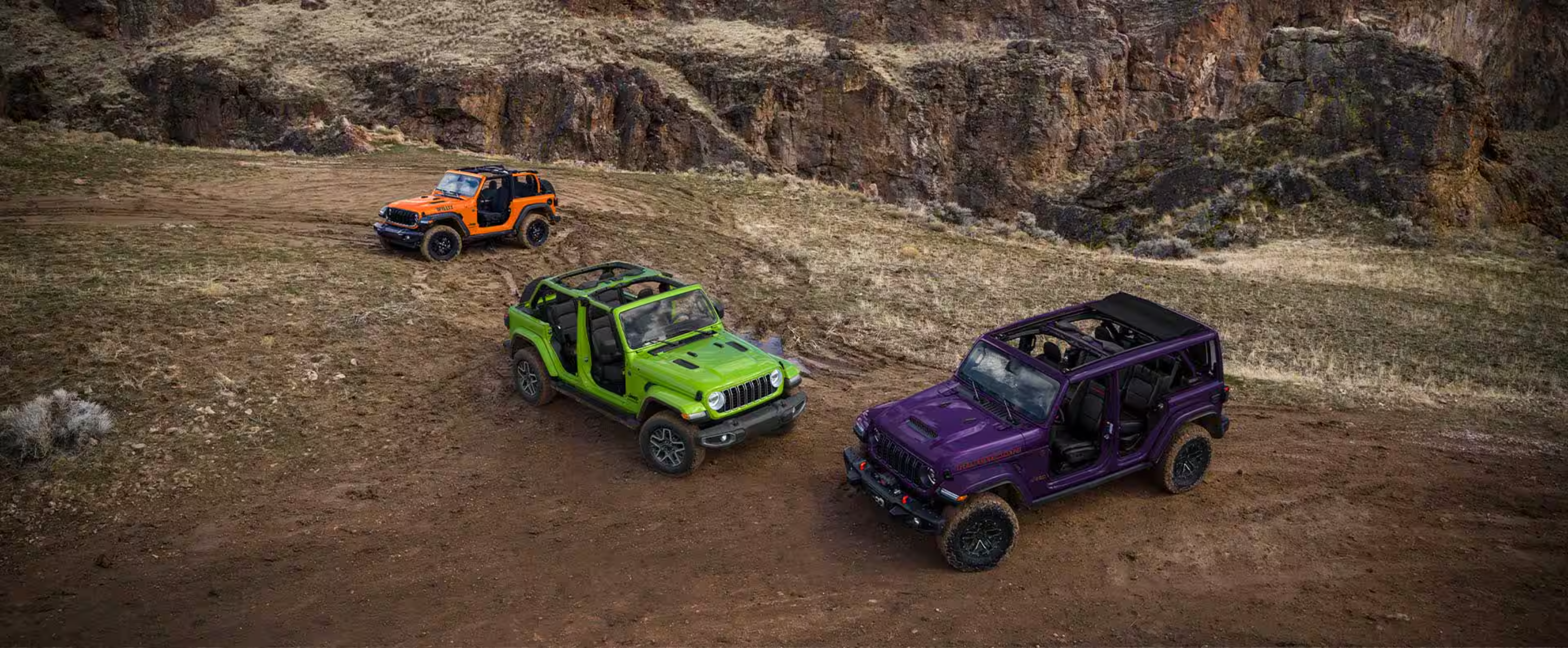 Three colorful SUVs in purple, green, and orange are parked on a dry, grassy plain against a backdrop of rugged rocky cliffs.