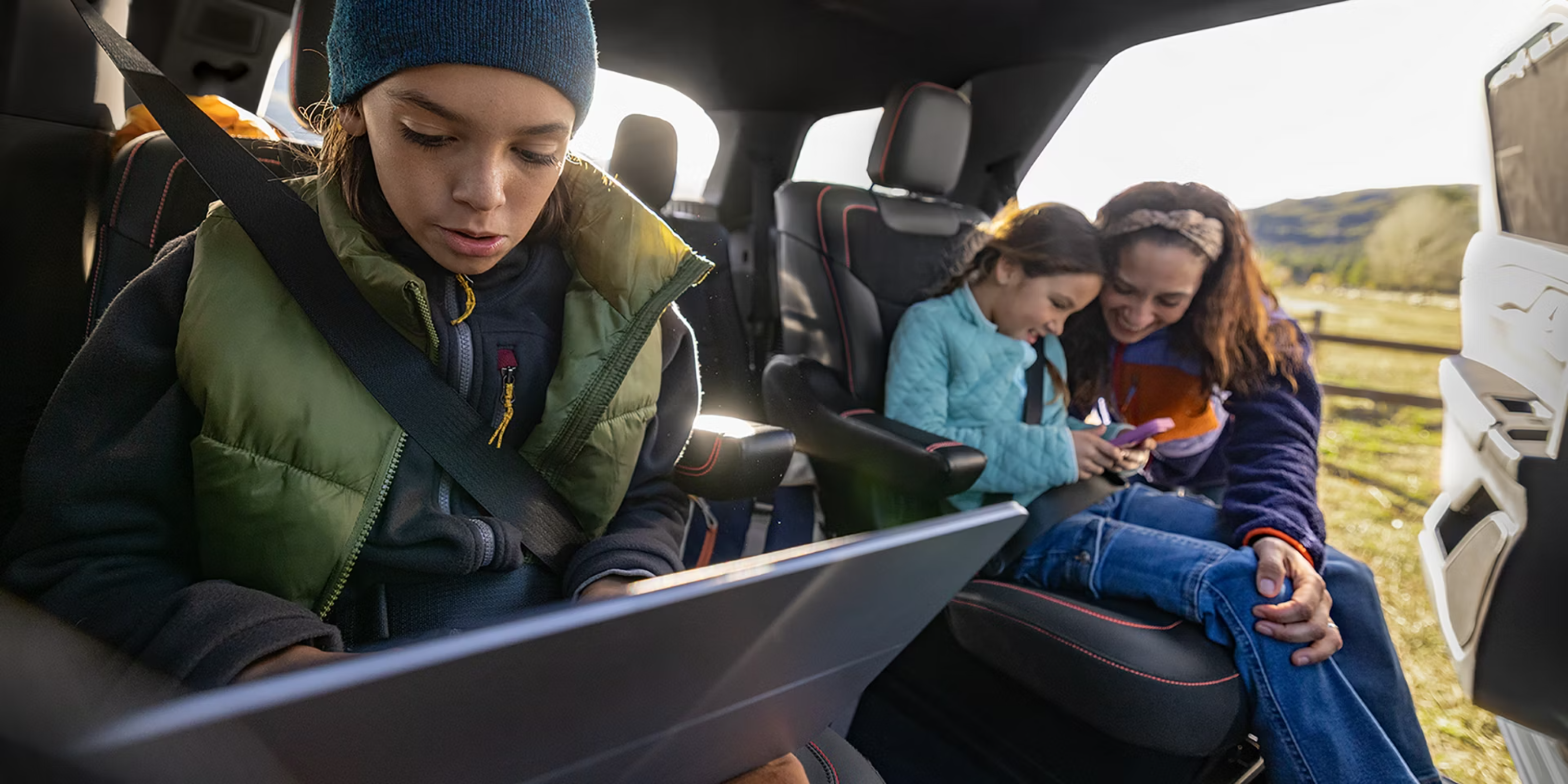 A young boy in a green vest looks at a tablet in the foreground, while a woman and girl smile together in the backseat against a sunny outdoor landscape.