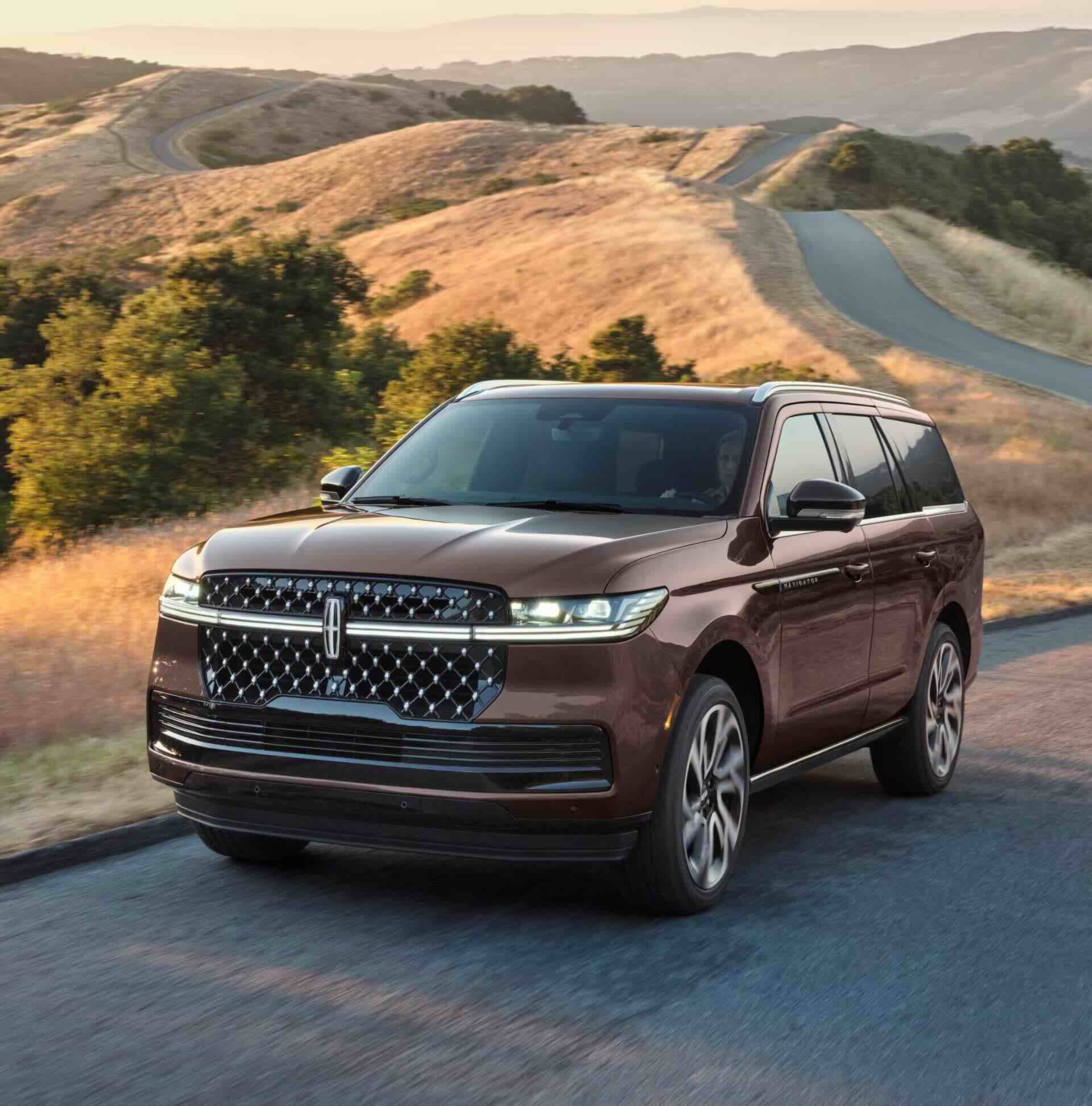A brown SUV drives on a paved road with rolling hills and a winding road in the background.