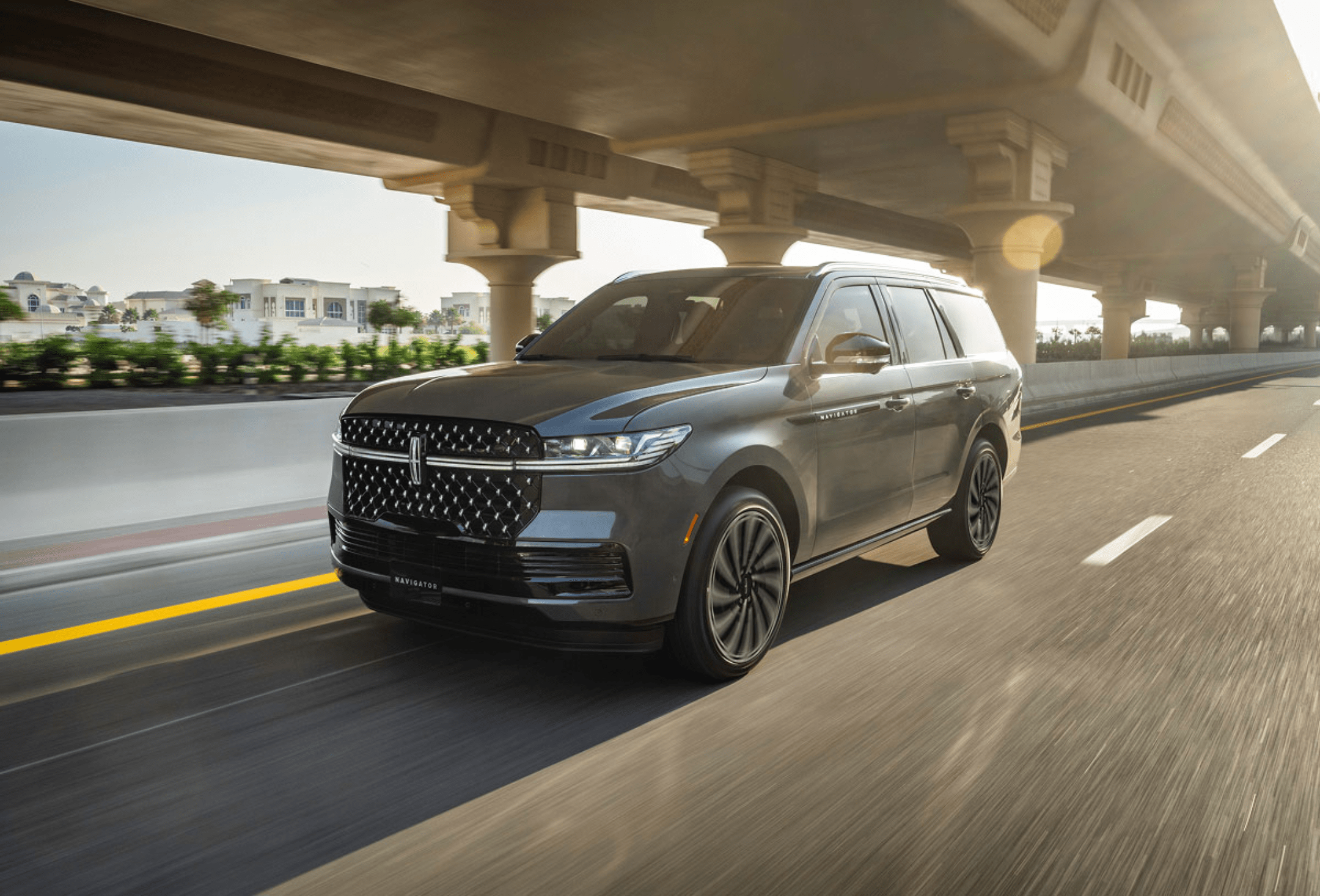 A dark gray SUV drives on a highway under a concrete overpass, with buildings and trees visible in the distance.