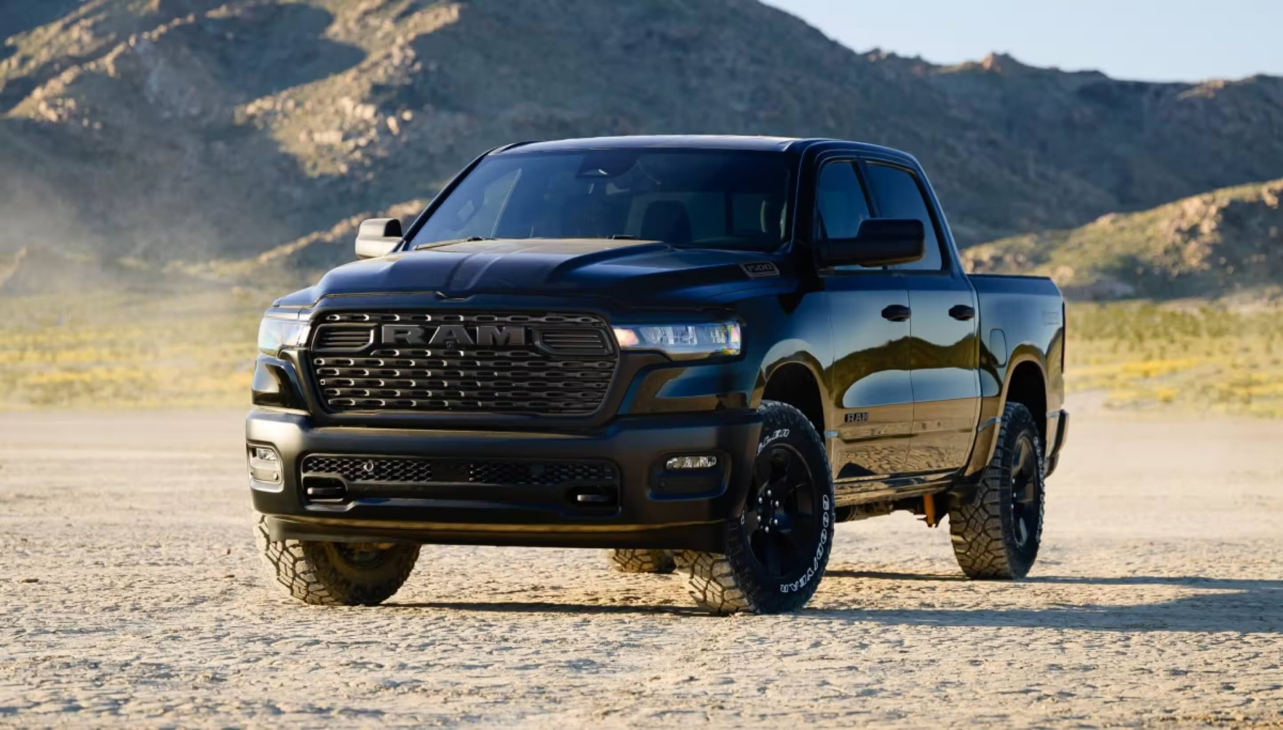 A black truck sits on sandy terrain with rolling hills and mountains in the background.