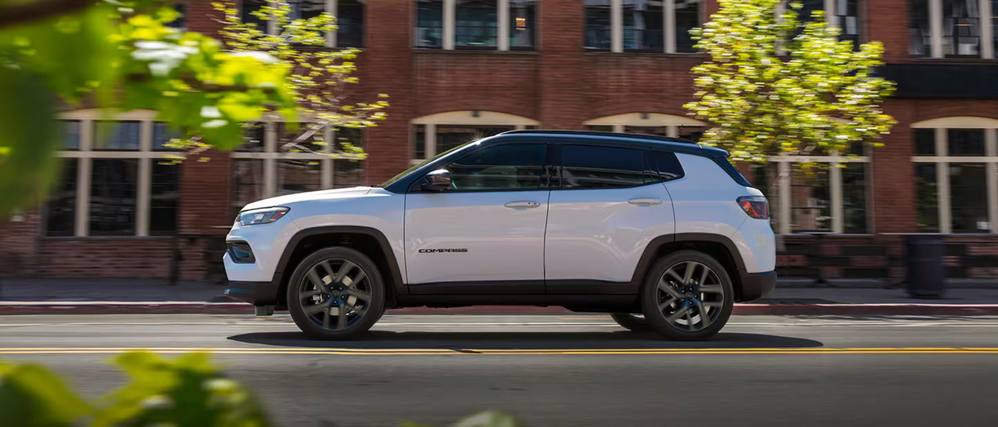 A white SUV drives along a city street with a red brick building and green foliage in the background.