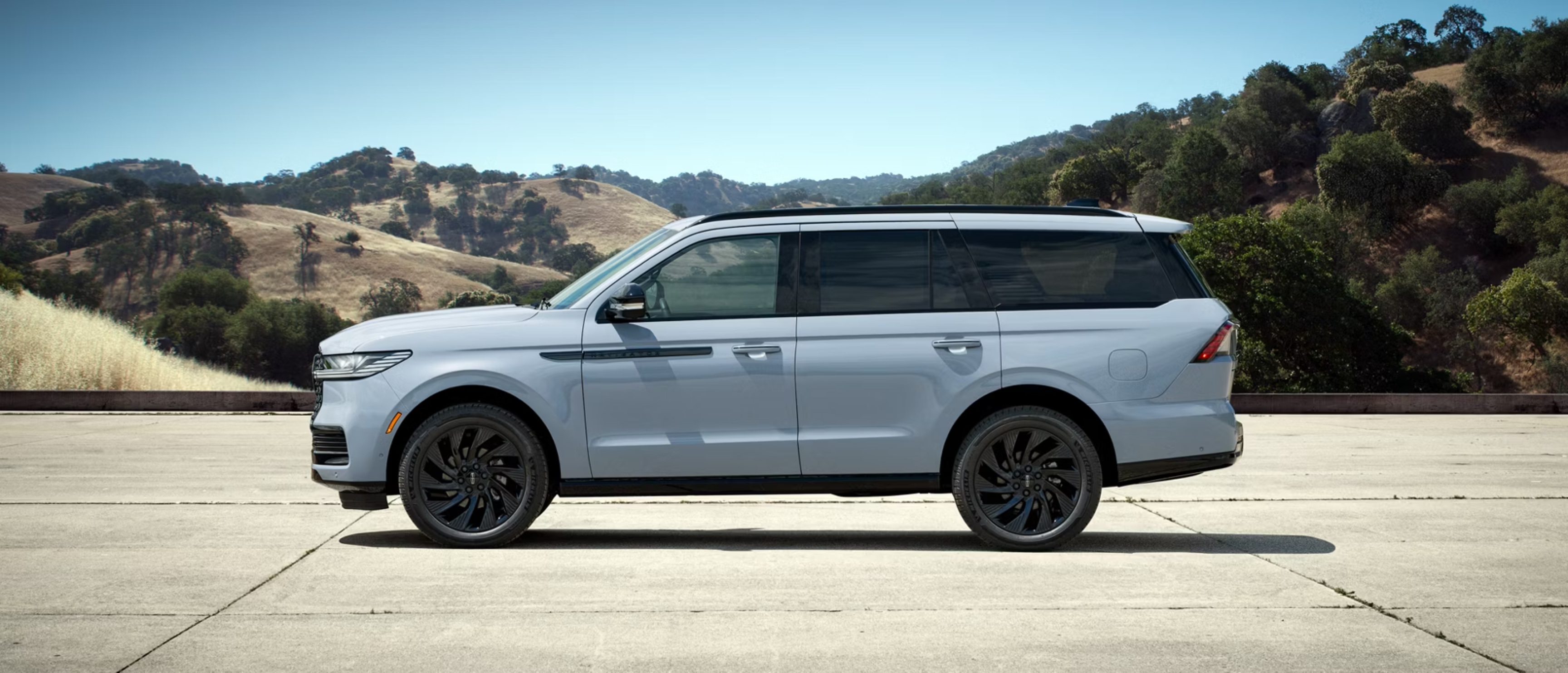 A light blue SUV with black wheels is parked on a concrete surface with rolling hills and trees in the background.