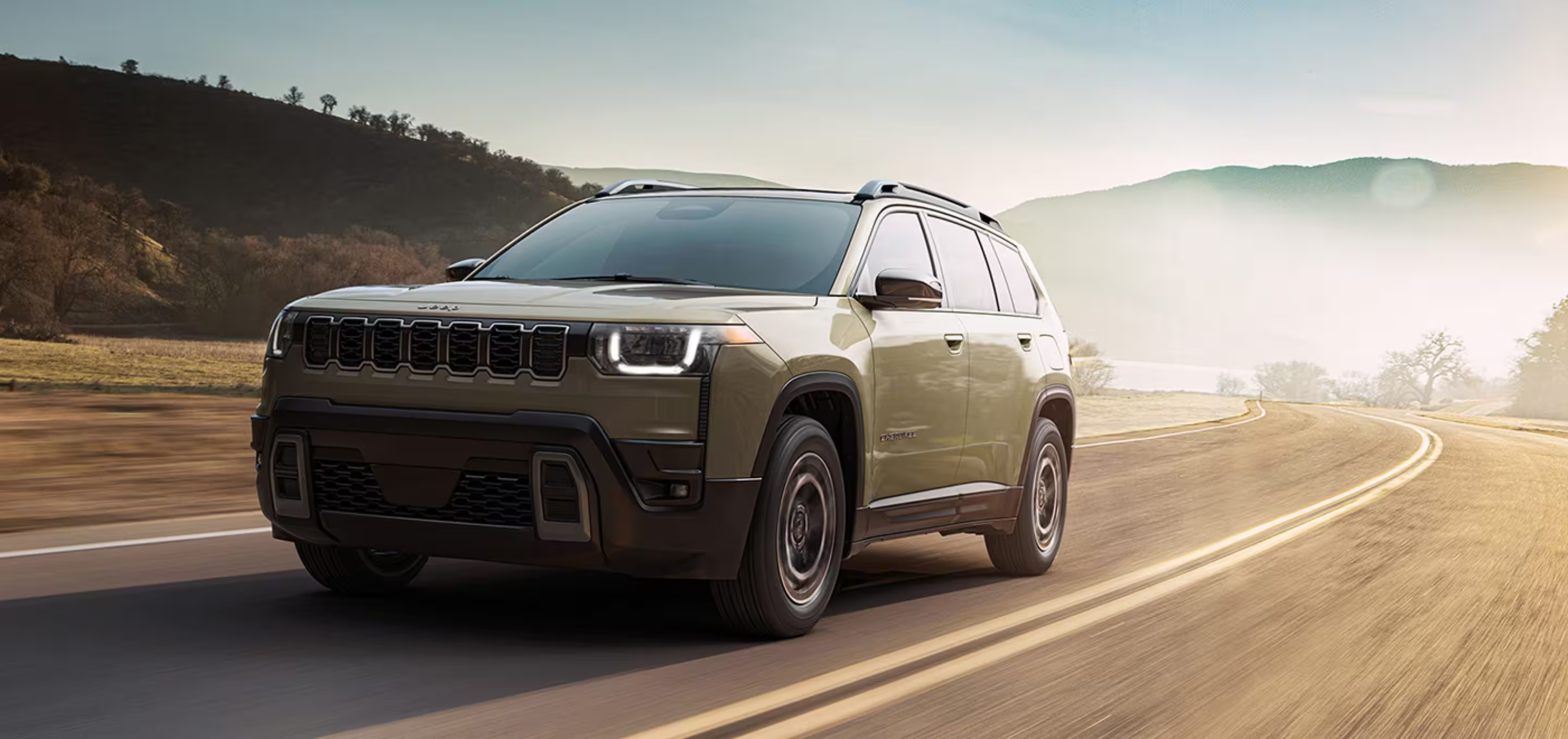 An olive green SUV drives along a winding road with hills and a hazy sky in the background.