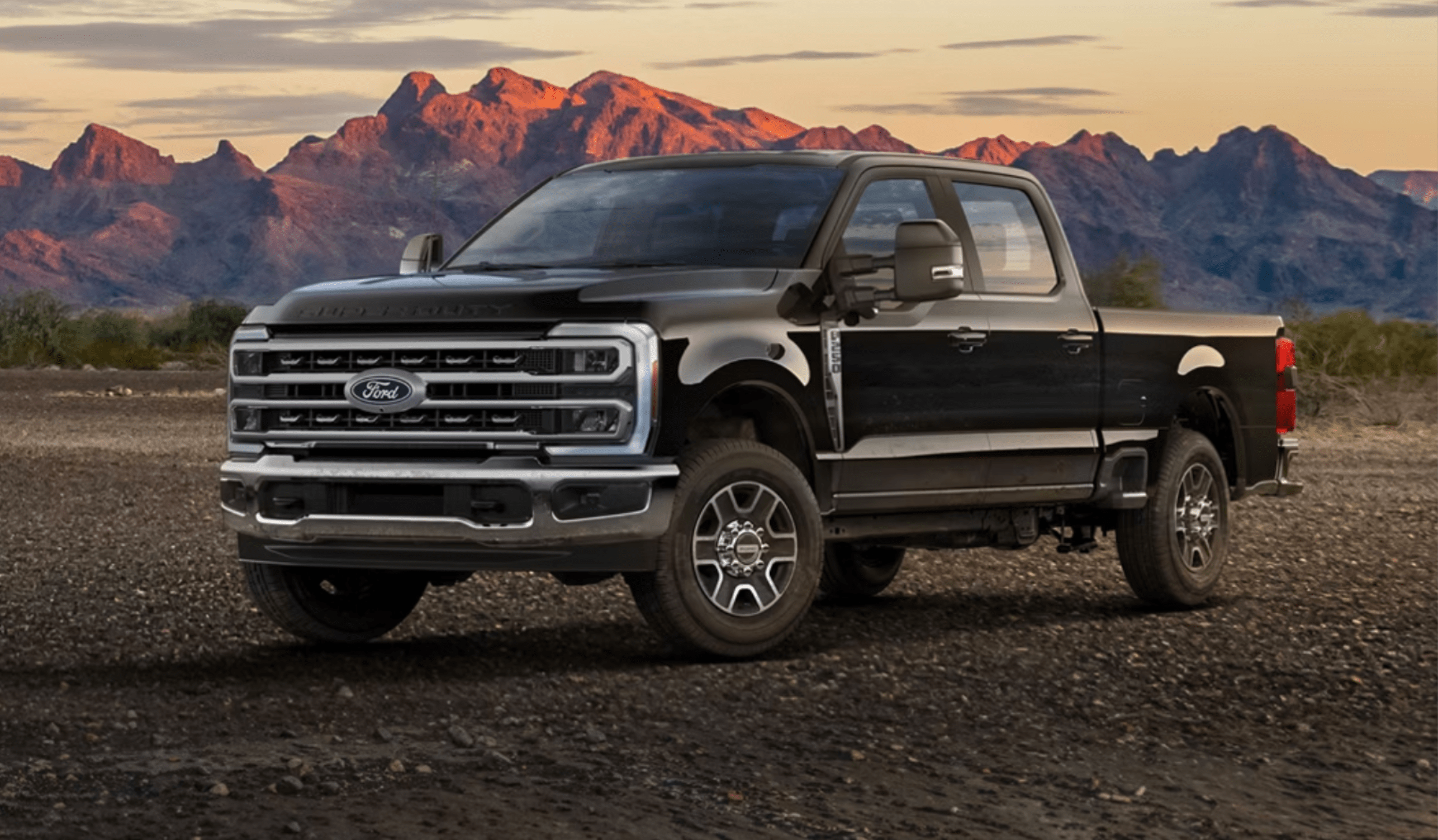 Black pickup truck on desert terrain with mountain landscape and sunset in the background.