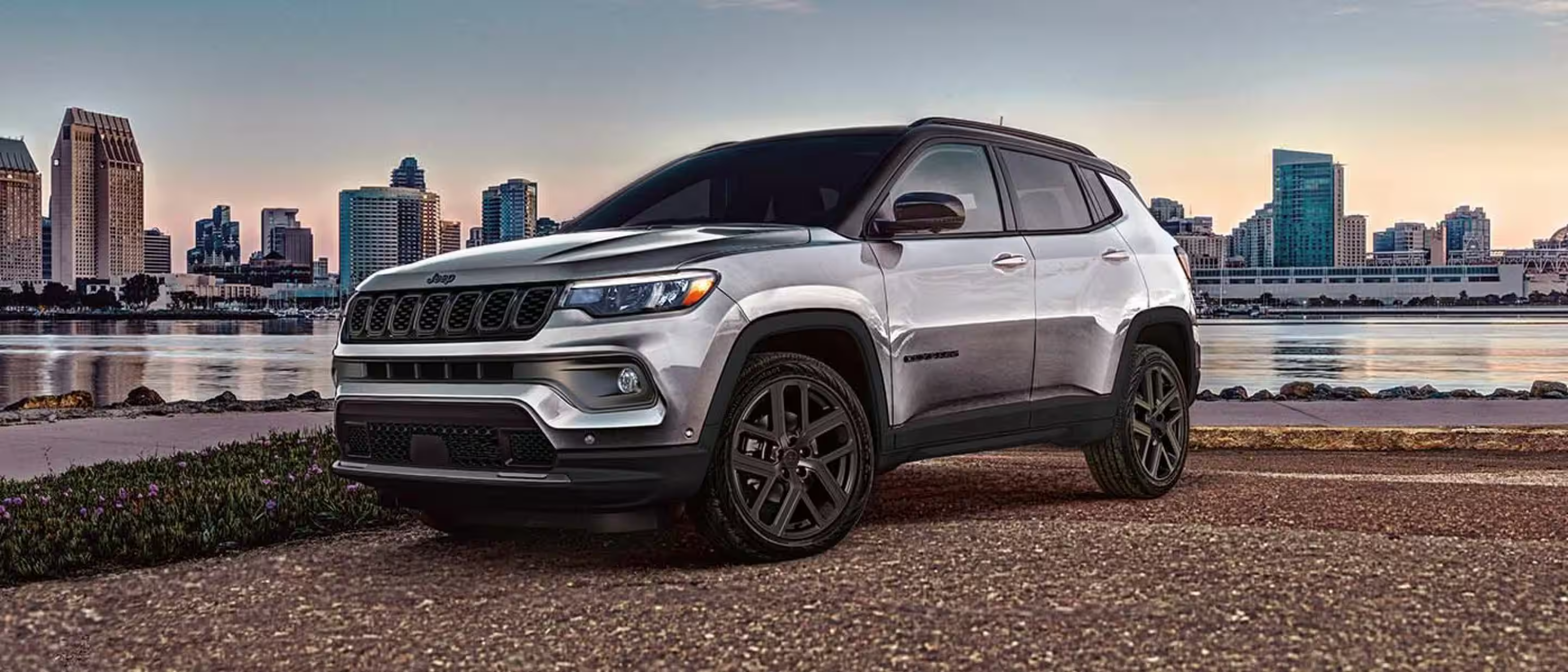 A silver SUV is parked on a paved area with a city skyline and waterfront in the background.