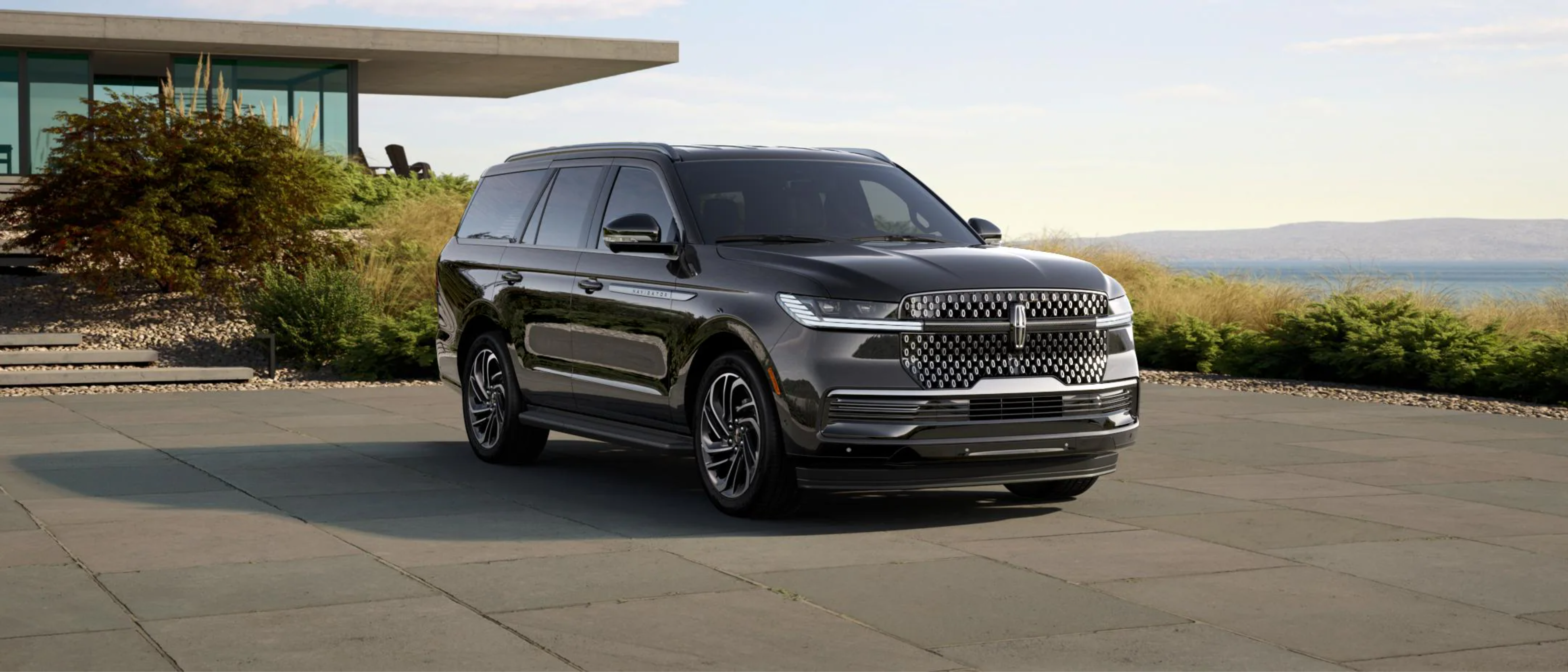 Dark gray SUV parked on paved driveway in front of modern house with large body of water and hills in background