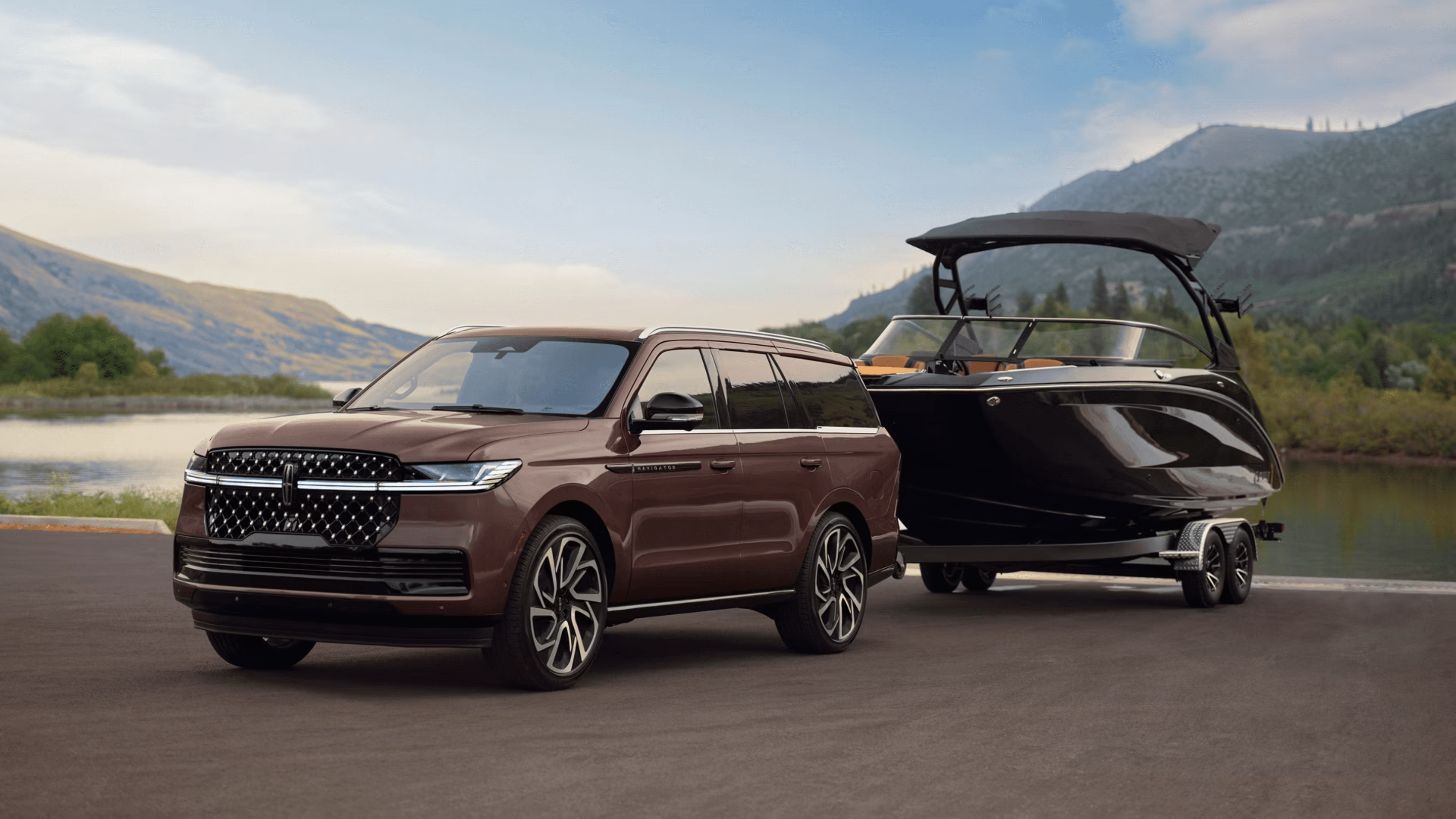 A brown SUV towing a sleek black motorboat on a trailer, set against a backdrop of calm water and distant mountains.