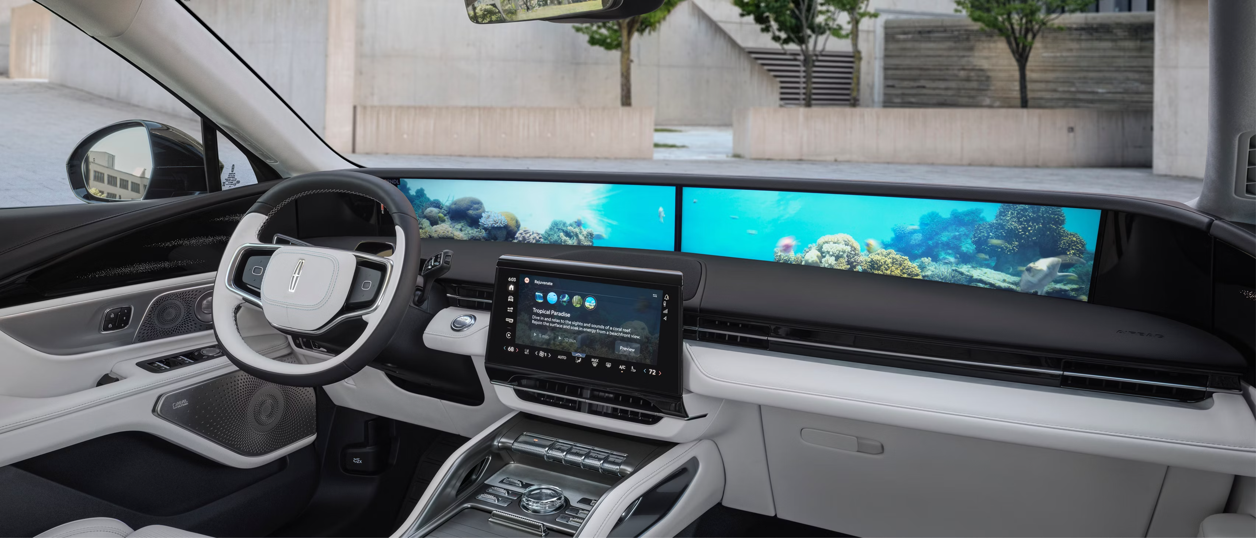 Modern black and white car interior with digital dashboard displaying ocean scene and concrete building visible through the windshield.