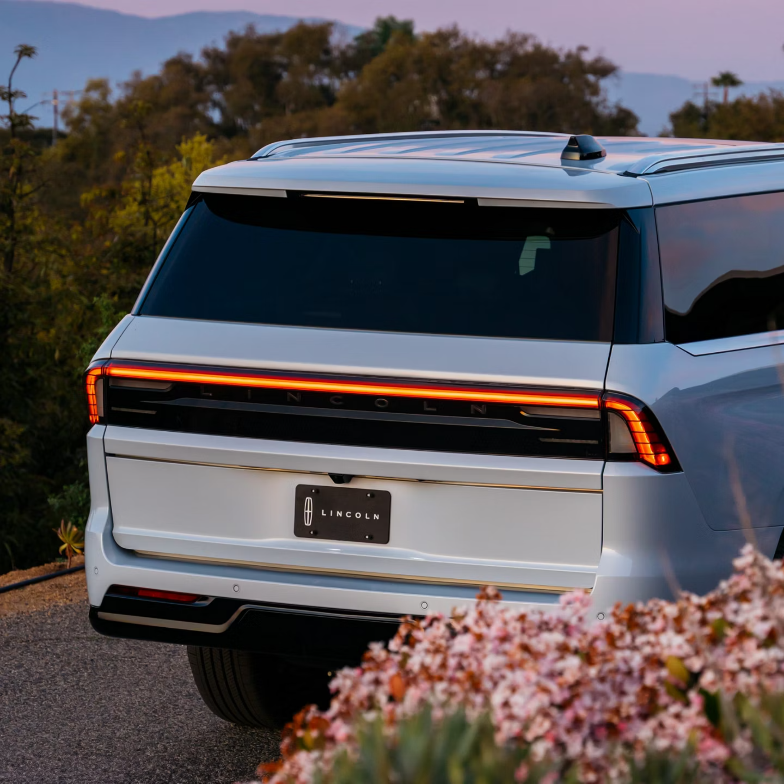 A white SUV is parked on a gravel driveway, with soft pink flowers in the foreground and trees and a dusky sky in the background.