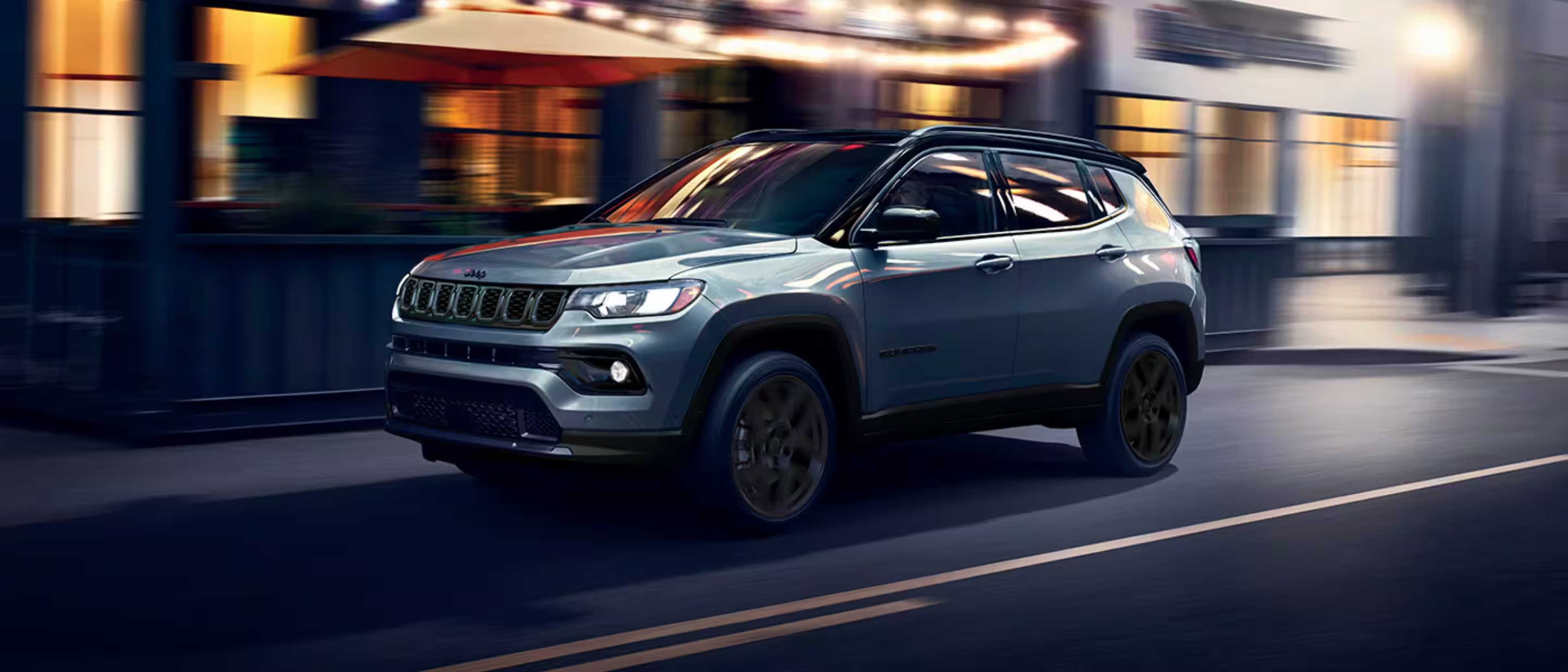 A silver SUV drives down a city street at night with blurred storefronts and warm lights in the background.