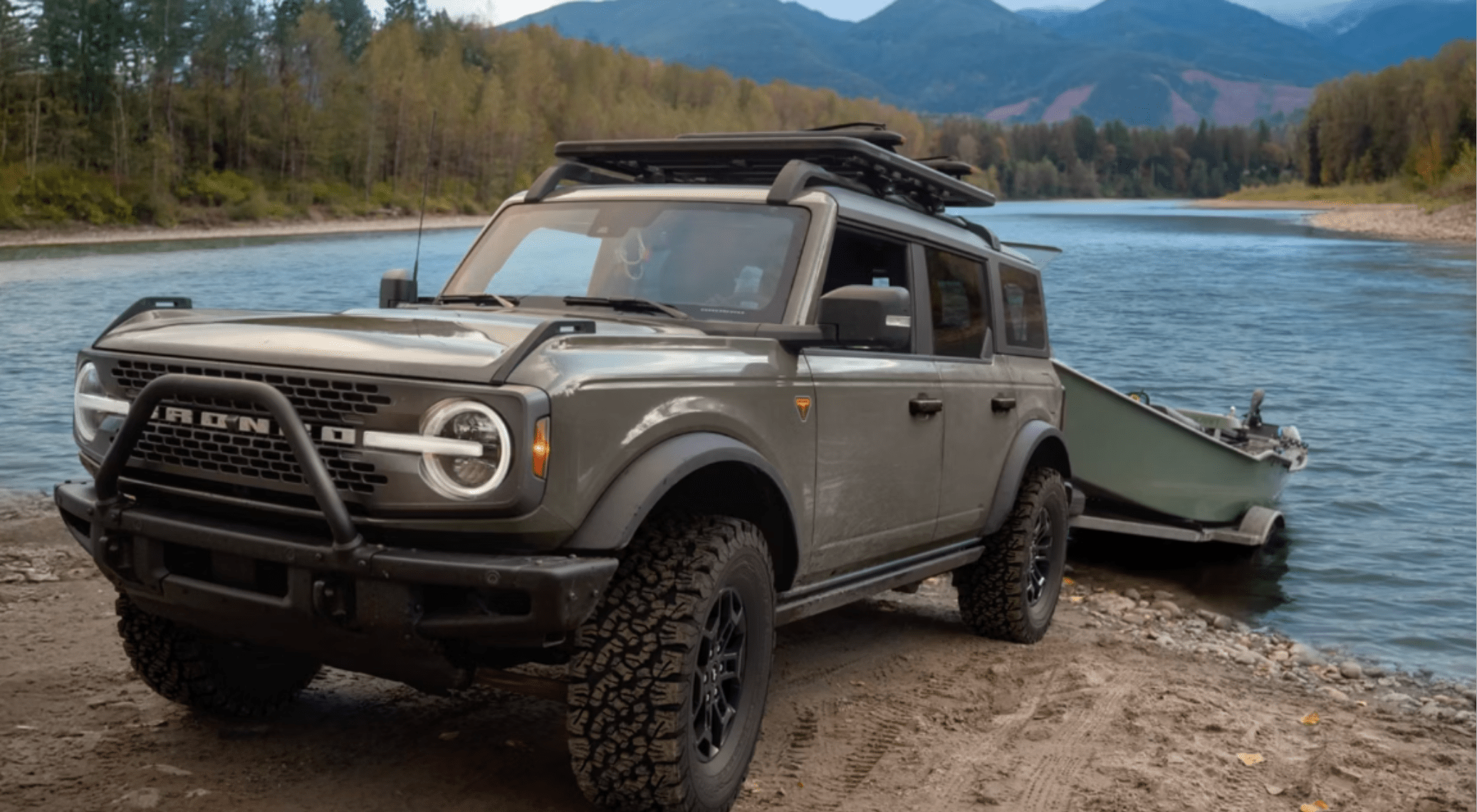 An SUV with a roof rack parked on a sandy shore next to a boat, with a calm river and forested mountains in the background.