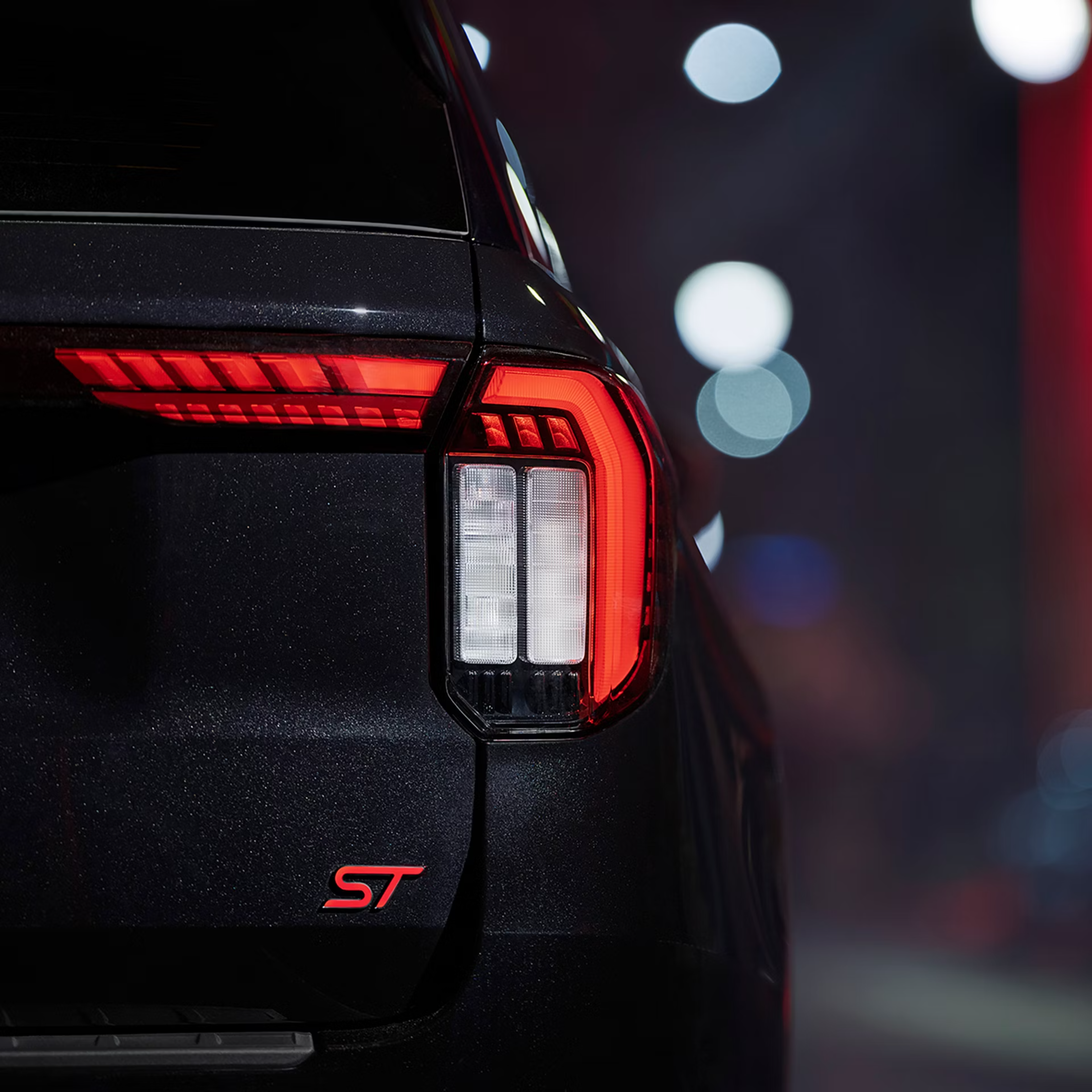 Close-up of a dark metallic car's rear taillight and red "ST" badge with city lights in the background.