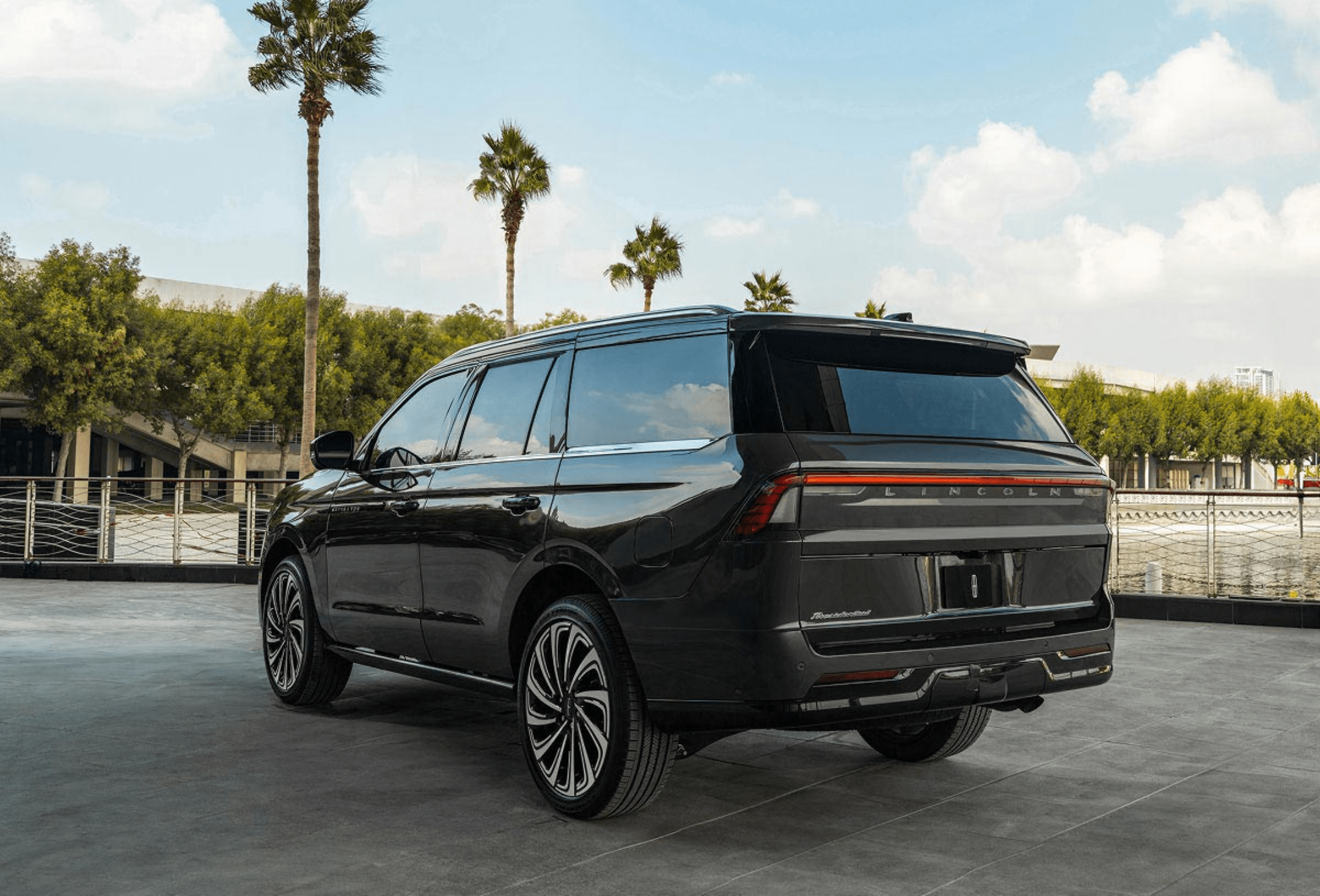 Black SUV with custom wheels parked on a paved lot with palm trees and a building in the background.